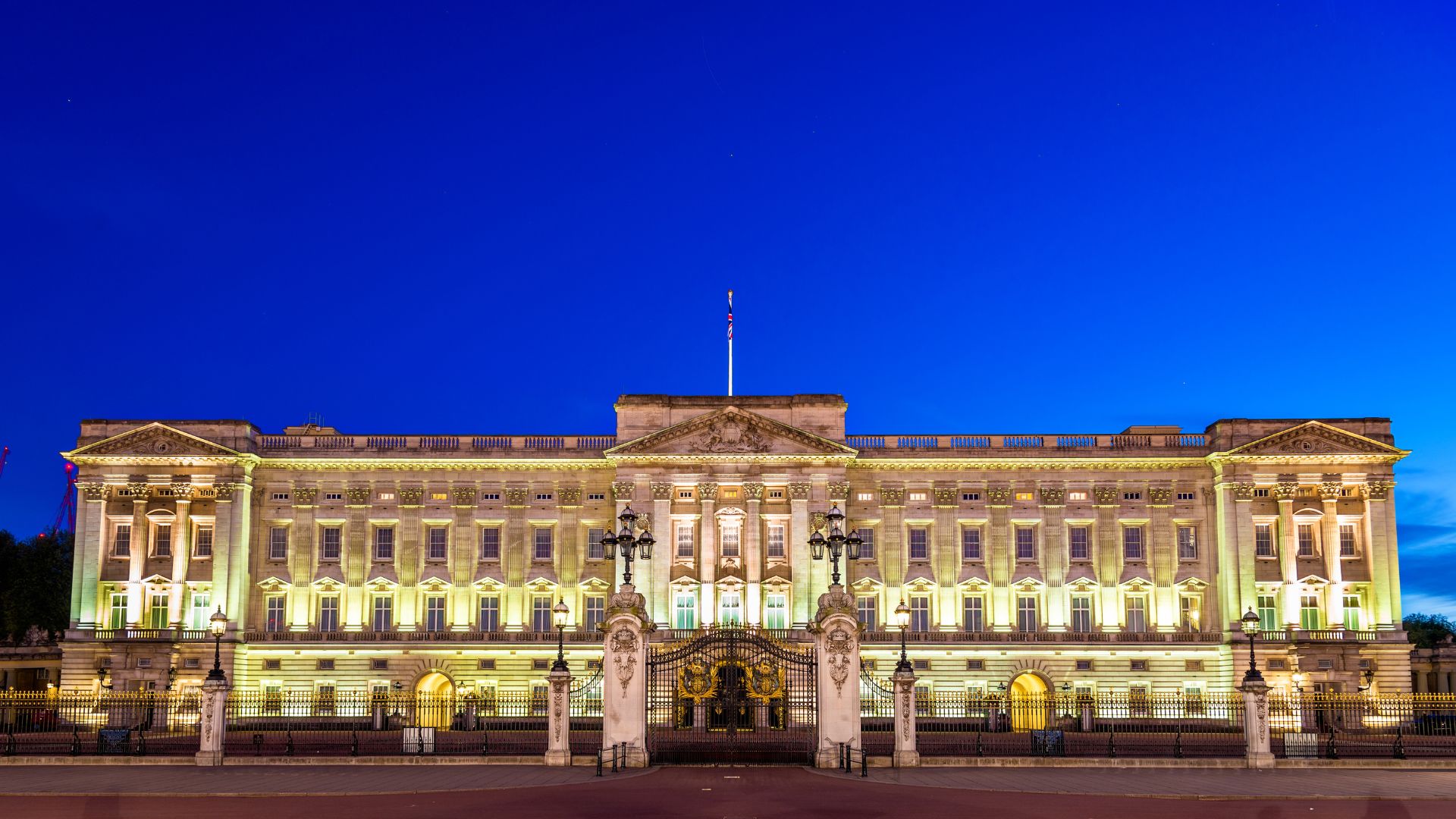 An evening shot of Buckingham Palace, illuminated with warm yellow lights against a deep blue twilight sky. The grand facade of the palace is visible, along with its prominent gates and surrounding lampposts.