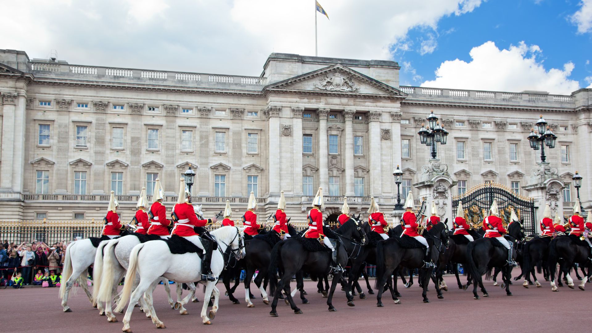 A wide shot of Buckingham Palace with its grand facade and flag flying, in front of which a procession of mounted Household Cavalry in red uniforms and bearskin hats are parading on both white and black horses.