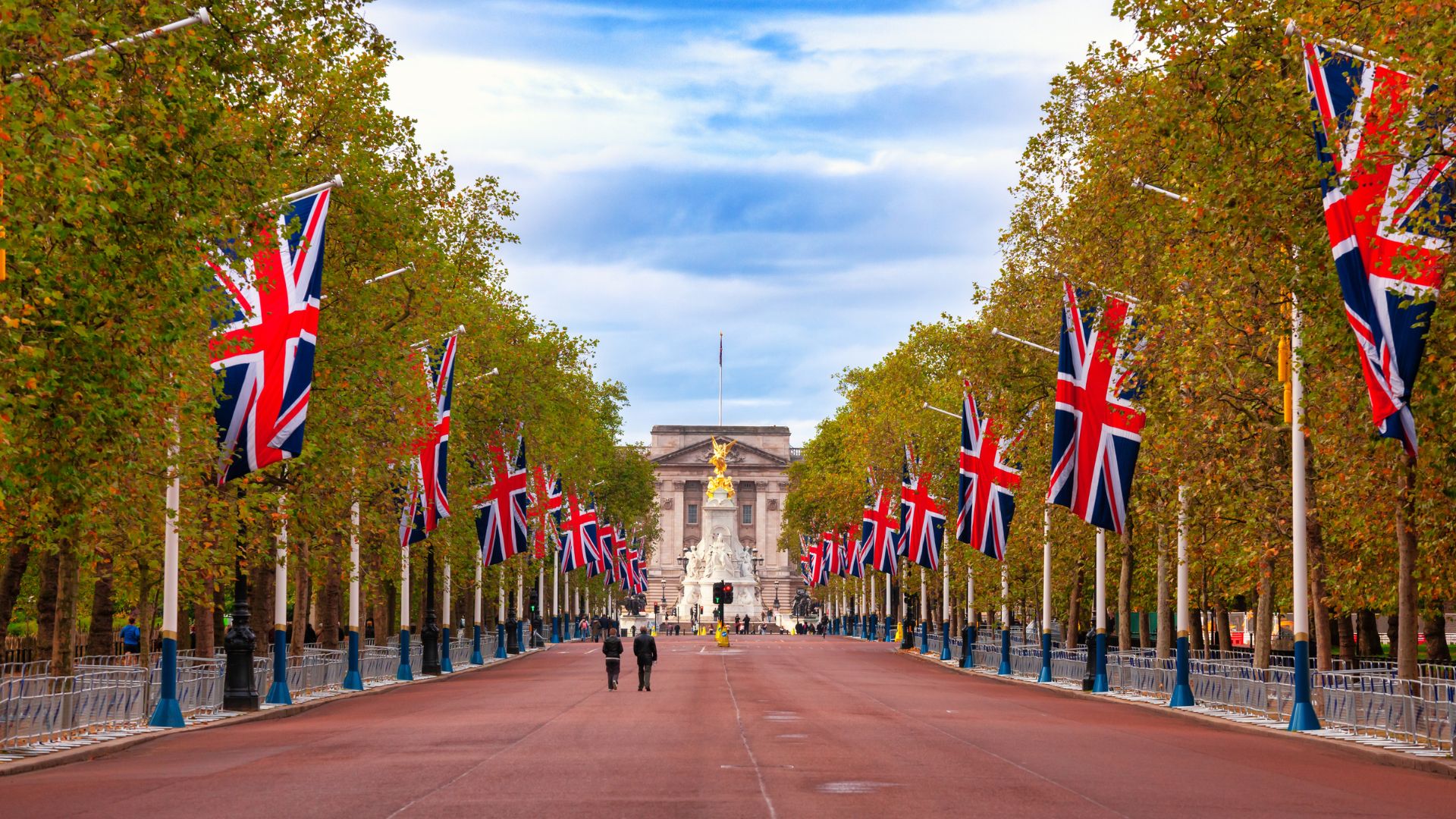 A wide, tree-lined avenue, The Mall, leads towards Buckingham Palace under a cloudy sky, with numerous Union Jack flags displayed prominently along both sides of the road. Two figures walk in the distance on the red-paved road.
