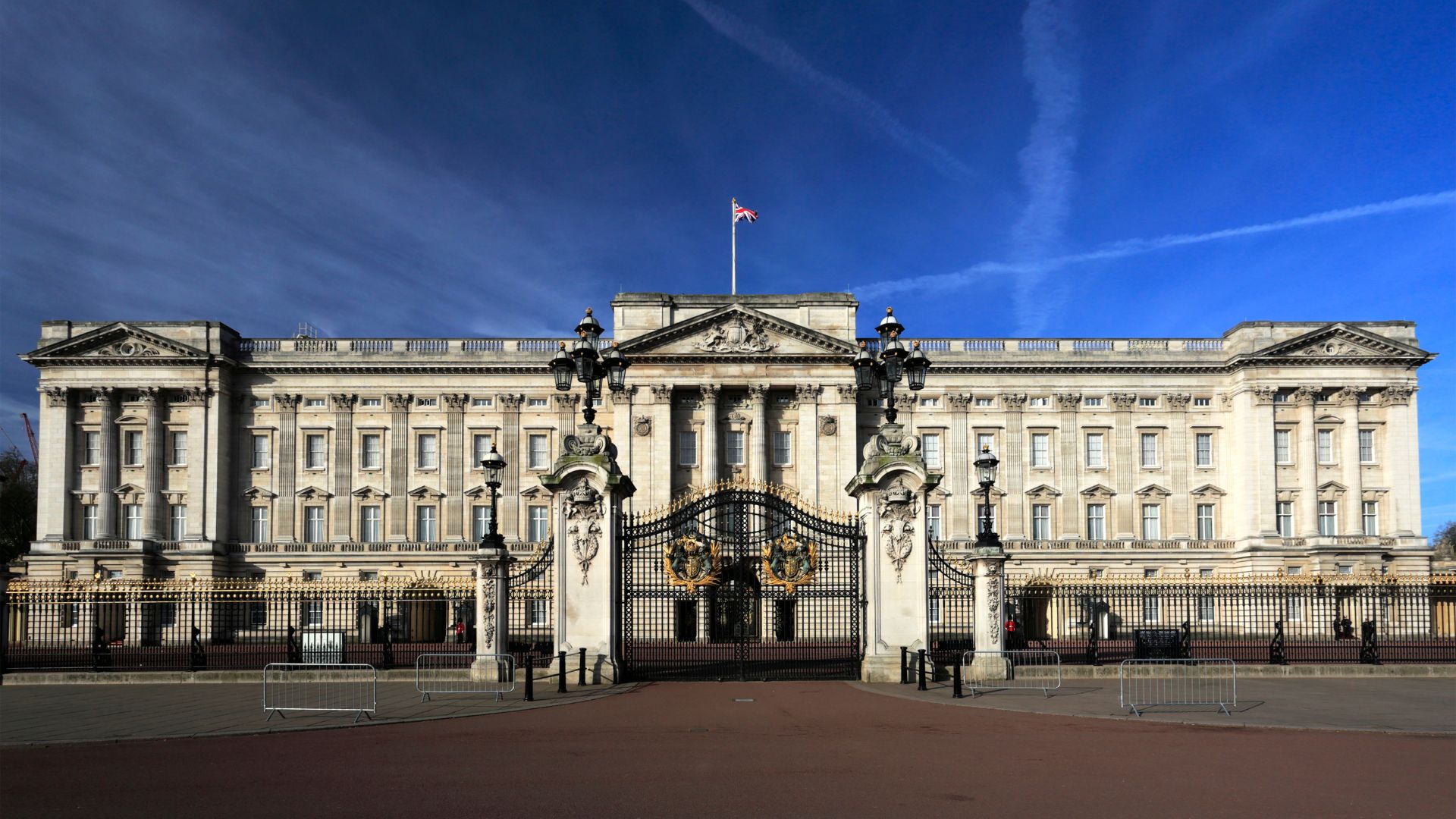 A wide shot of Buckingham Palace, showcasing its grand facade, ornate gates, and the Union Flag flying atop the building under a clear blue sky.
