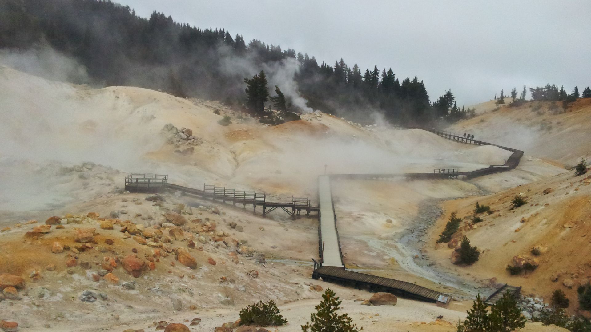 A wide-angle view of Bumpass Hell, a hydrothermal area in Lassen Volcanic National Park, California, featuring a winding wooden boardwalk through steaming fumaroles and colorful, mineral-stained ground, with a forested hillside in the background under a cloudy sky.