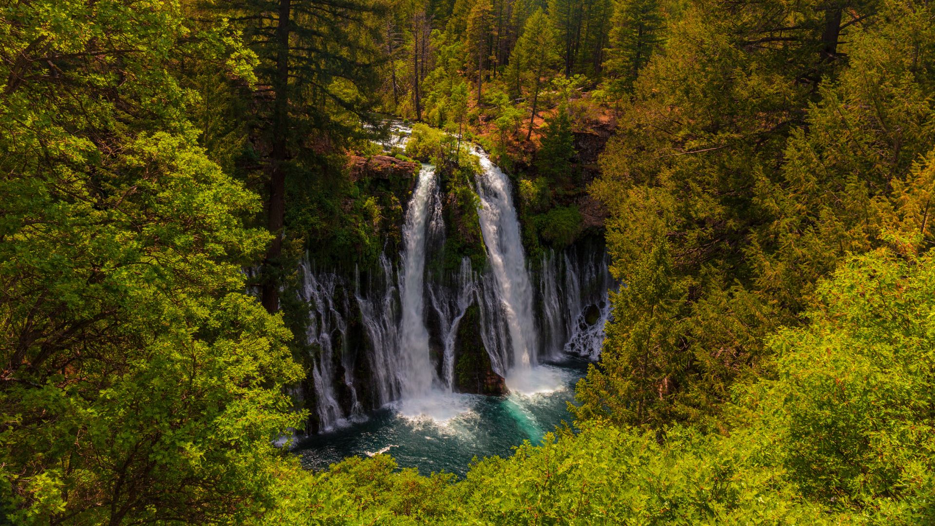 A wide waterfall cascades over a lush, moss-covered cliff face, with tall evergreen trees visible above and a pool of water at the base, under a partly cloudy sky.