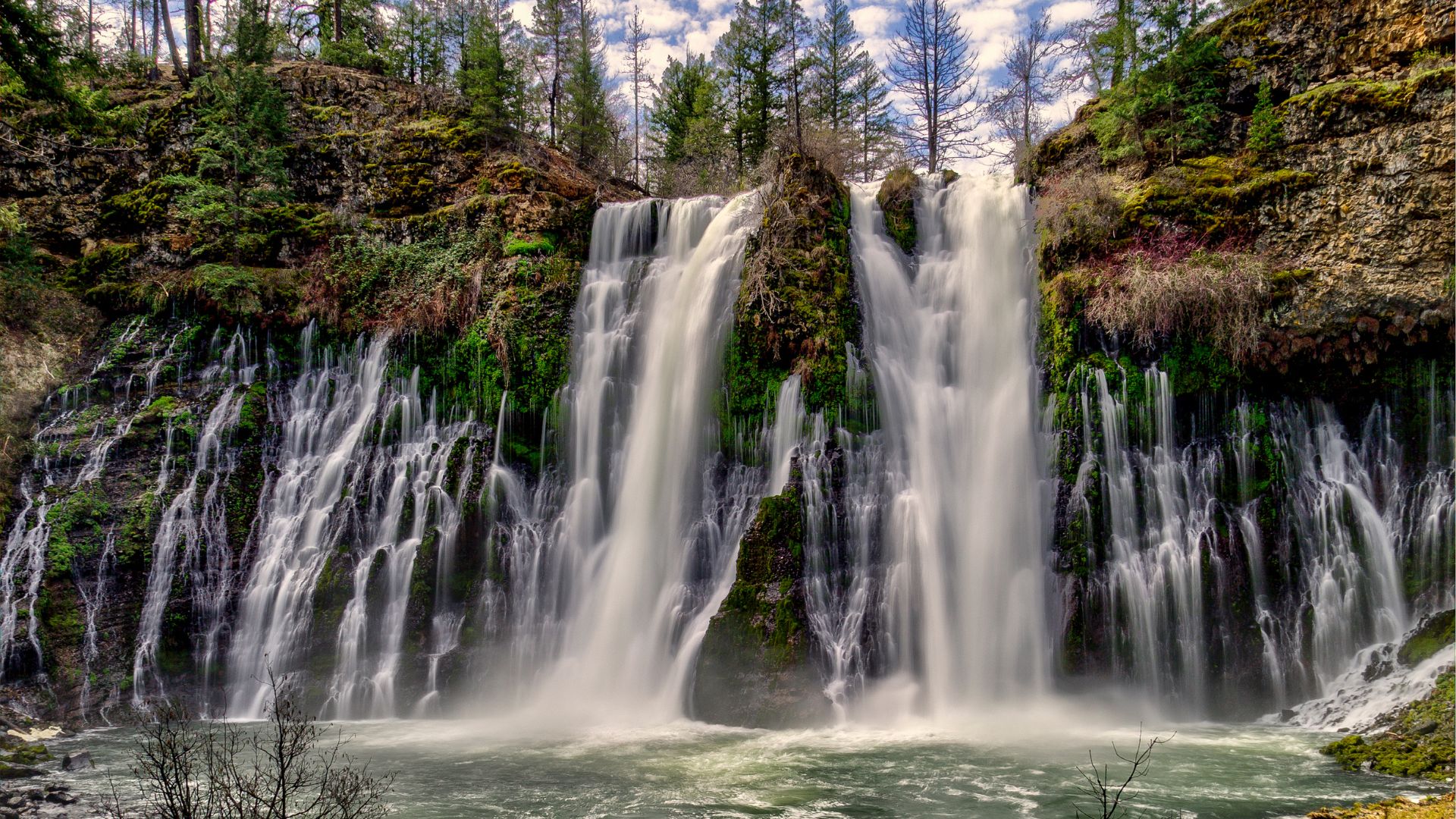 A wide waterfall cascades over a lush, moss-covered cliff face, with tall evergreen trees visible above and a pool of water at the base, under a partly cloudy sky.