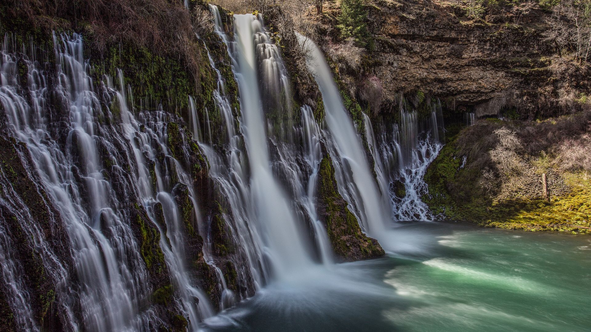 A wide-angle shot of Burney Falls, a large waterfall cascading over a moss-covered cliff face into a turquoise pool below, surrounded by lush vegetation and rocky terrain.