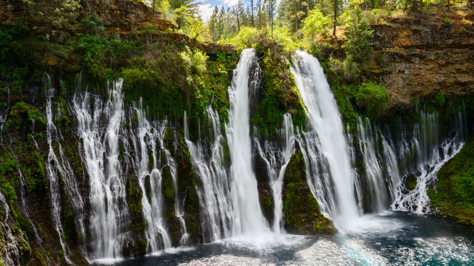 A wide shot of Burney Falls, a large, multi-tiered waterfall cascading over a mossy cliff face into a clear blue pool at its base, surrounded by lush green trees and rocky terrain.
