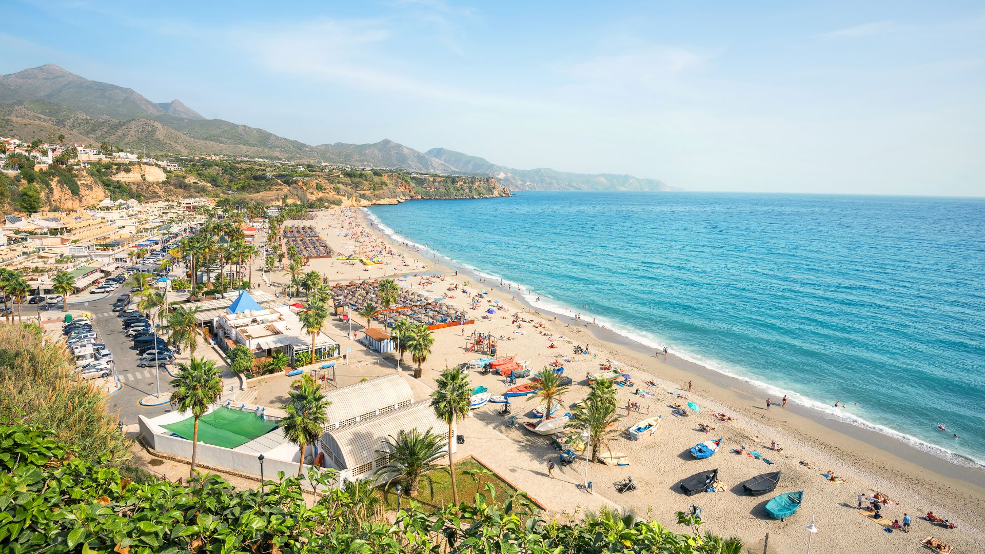 An aerial view of a bustling beach in Nerja, Spain, with turquoise waters, sandy shores dotted with umbrellas and boats, and a town built into the cliffs with buildings and palm trees, all backed by mountains under a clear sky.