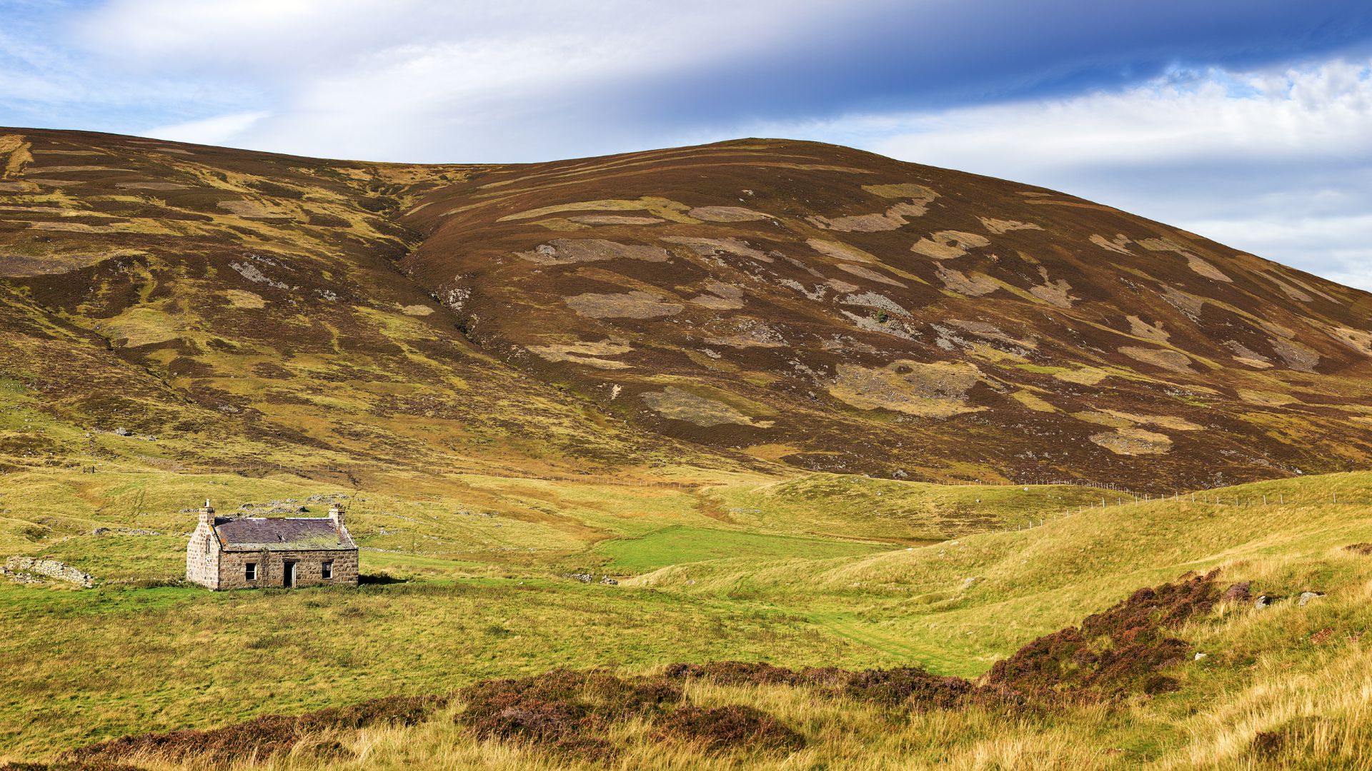 An abandoned stone house with a partially collapsed roof sits in a vast, green and brown mountainous landscape under a cloudy sky in the Scottish Highlands.