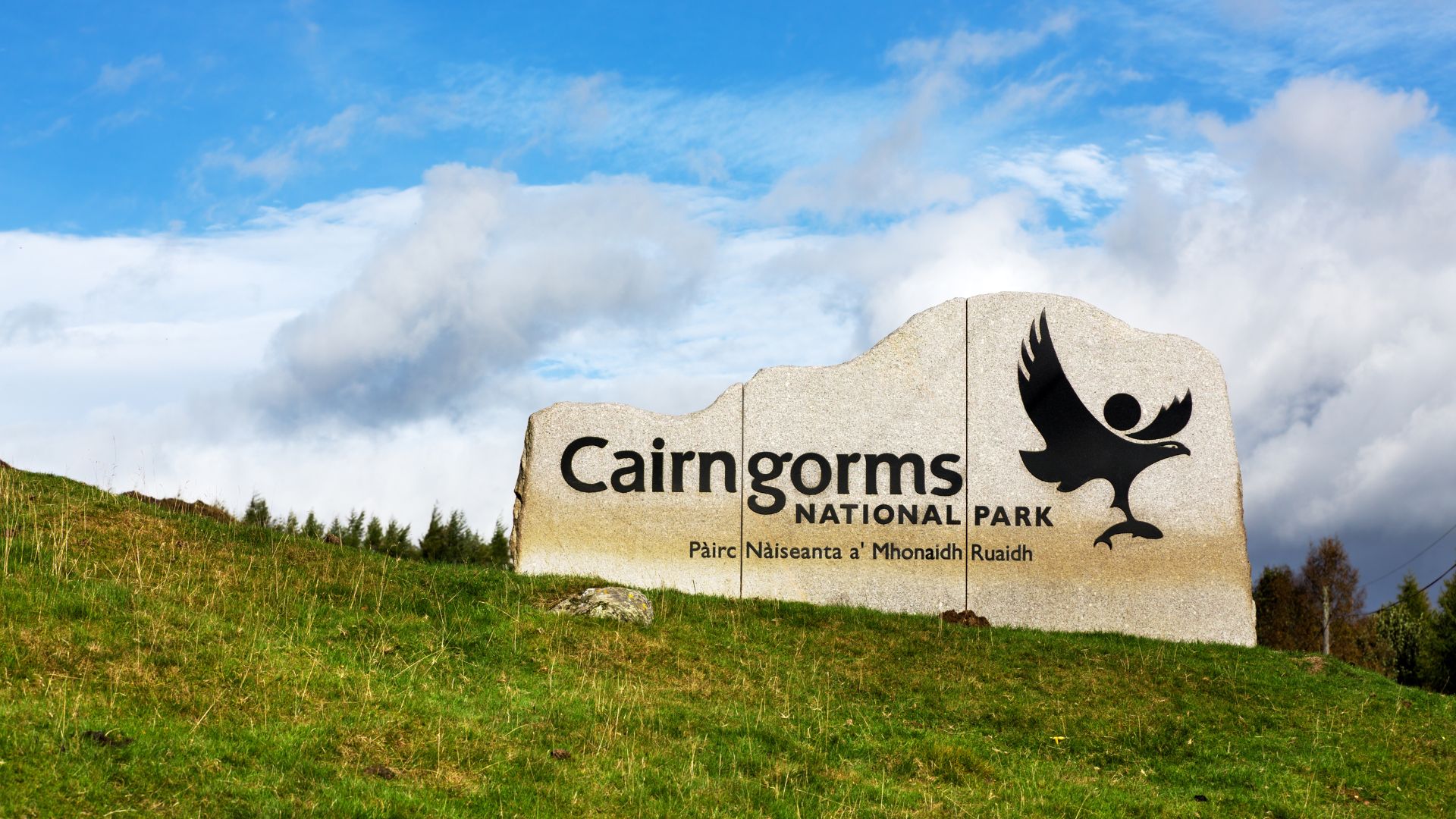 A wide shot of a large, rough-hewn stone sign for "Cairngorms NATIONAL PARK" on a grassy hillside under a cloudy blue sky, featuring a stylized bird logo on the right side of the sign.