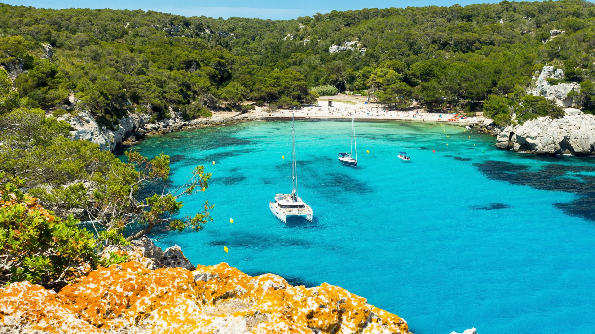 A high-angle view of Cala Macarella, a picturesque cove with strikingly clear turquoise water, white sand, and surrounded by lush green pine forests and rocky cliffs, with a few boats anchored in the calm waters.