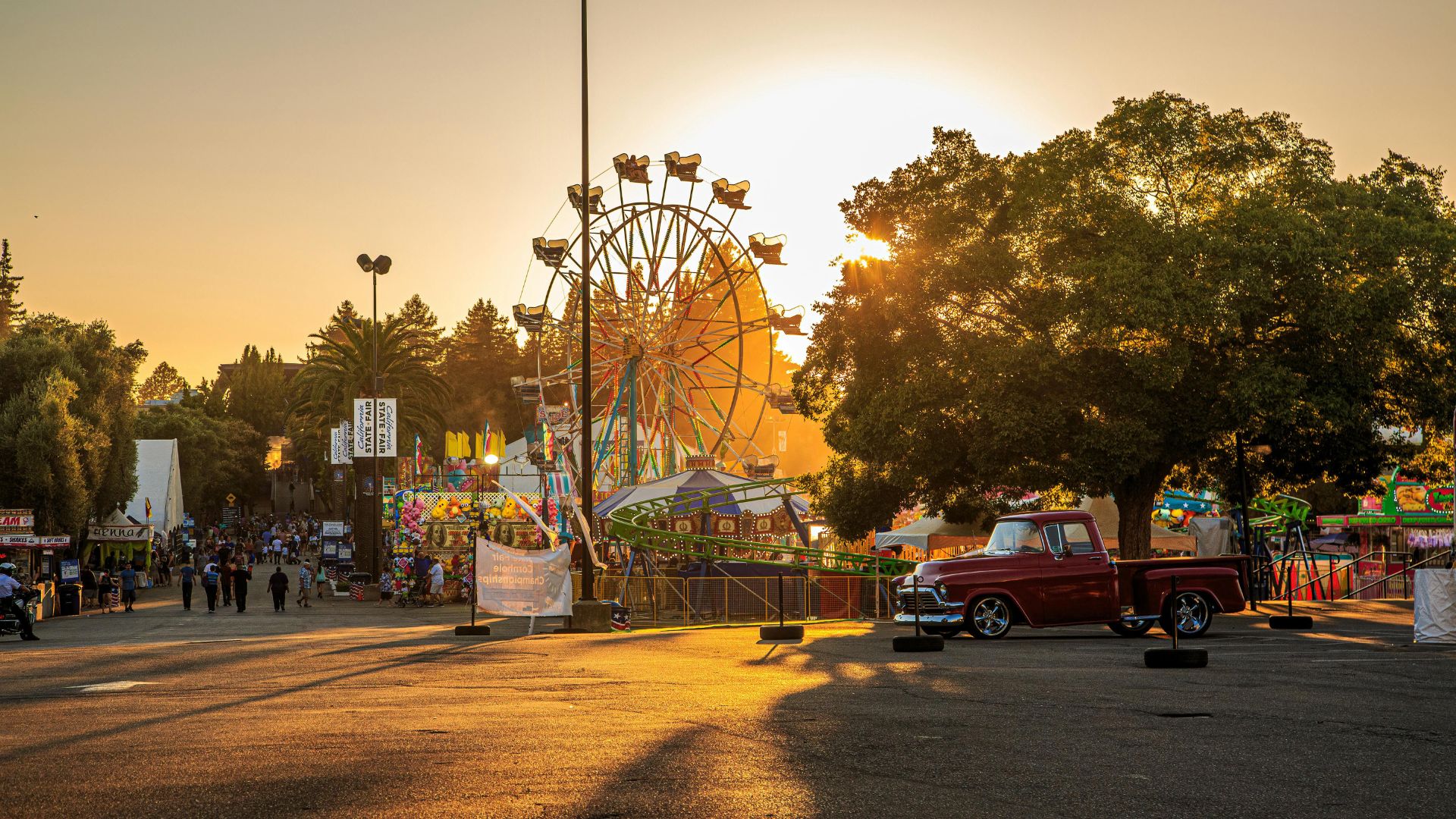 A vibrant sunset scene at a state fair, featuring a large Ferris wheel and other carnival rides illuminated against the golden sky, with a vintage red pickup truck parked in the foreground and fairgoers visible in the background.