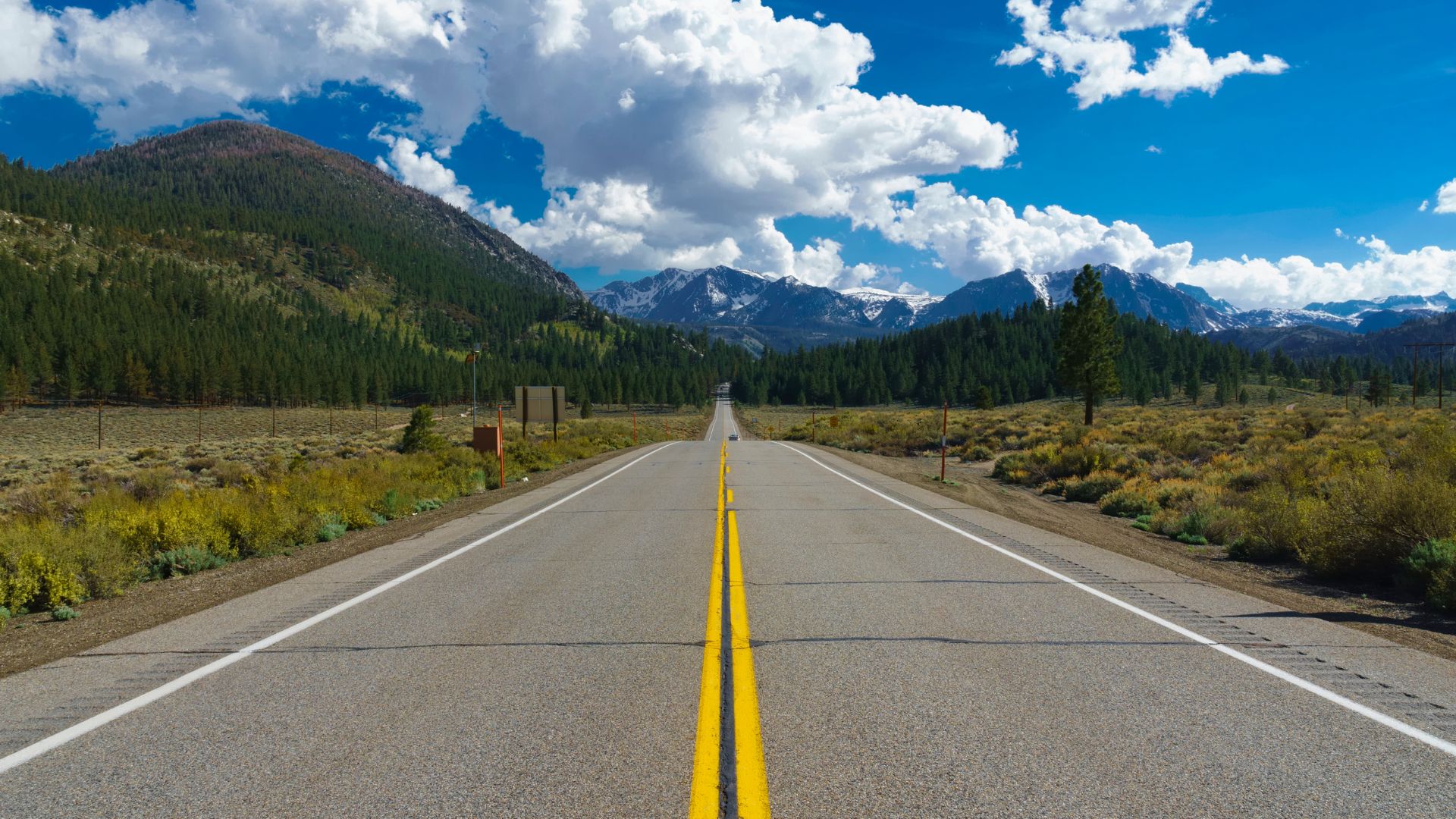 A long, straight two-lane highway, the June Lake Loop, stretches into the distance, framed by forested mountains and a clear blue sky with white clouds in the Eastern Sierra Nevada mountains of California. 