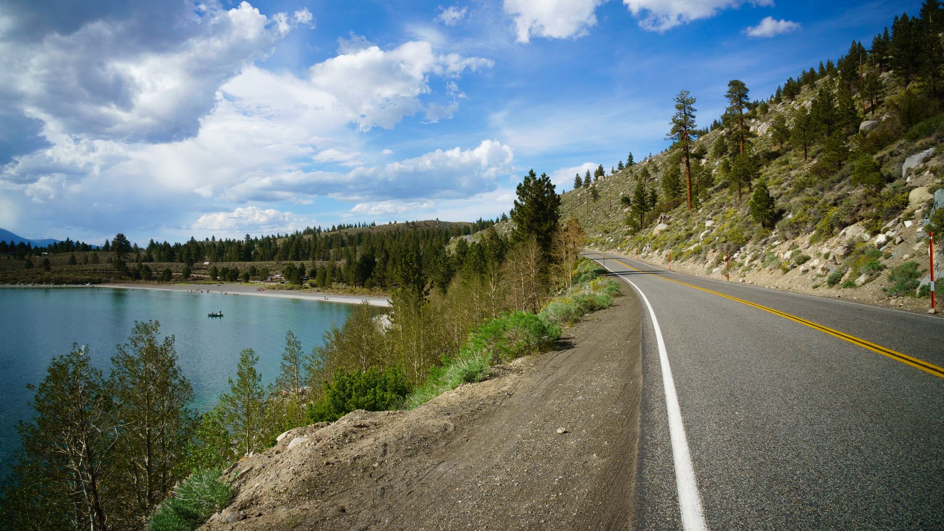 A scenic view of California State Route 158 (June Lake Loop) winding alongside a tranquil lake with forested hills and mountains under a partly cloudy sky.