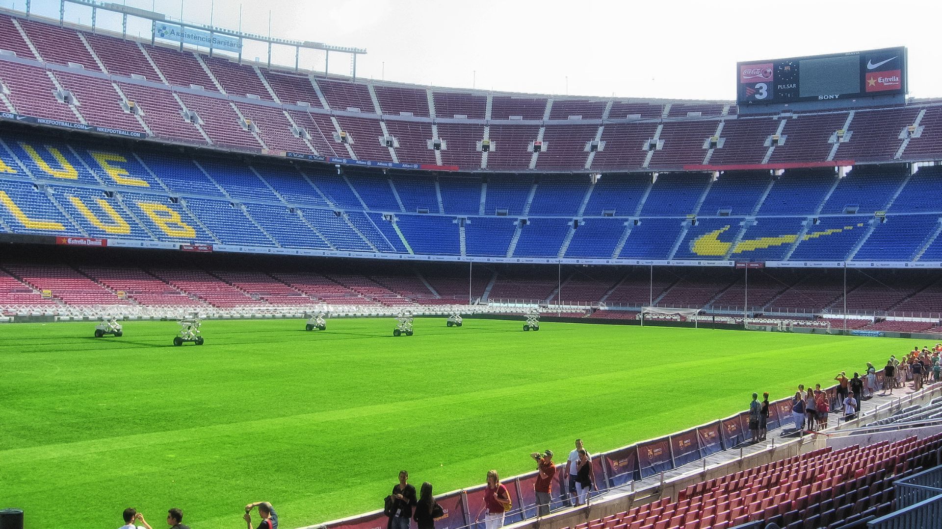 A wide-angle view of the interior of a large, empty football stadium, showing rows of red and blue seating, a bright green pitch with ground maintenance equipment, and a large scoreboard displaying "3" and a team logo. A few people are visible in the stands on the lower right. 