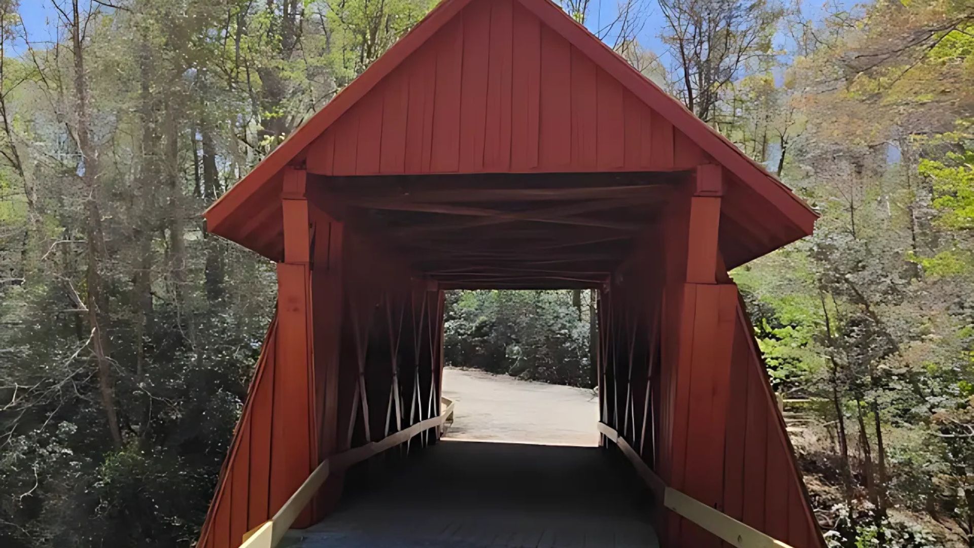 A red wooden covered bridge, Campbell's Covered Bridge, spans a small creek and leads to a gravel path, surrounded by lush green trees under a clear blue sky.