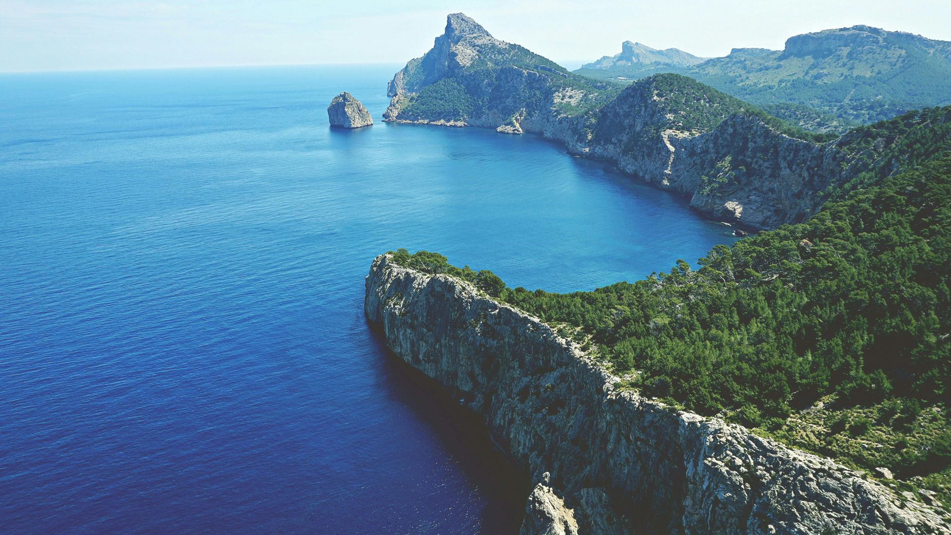 An aerial view of the dramatic, rugged coastline of Cap de Formentor, Majorca, featuring steep, forested cliffs dropping into the deep blue Mediterranean Sea, with a prominent rock formation (Es Colomer) in the distance.