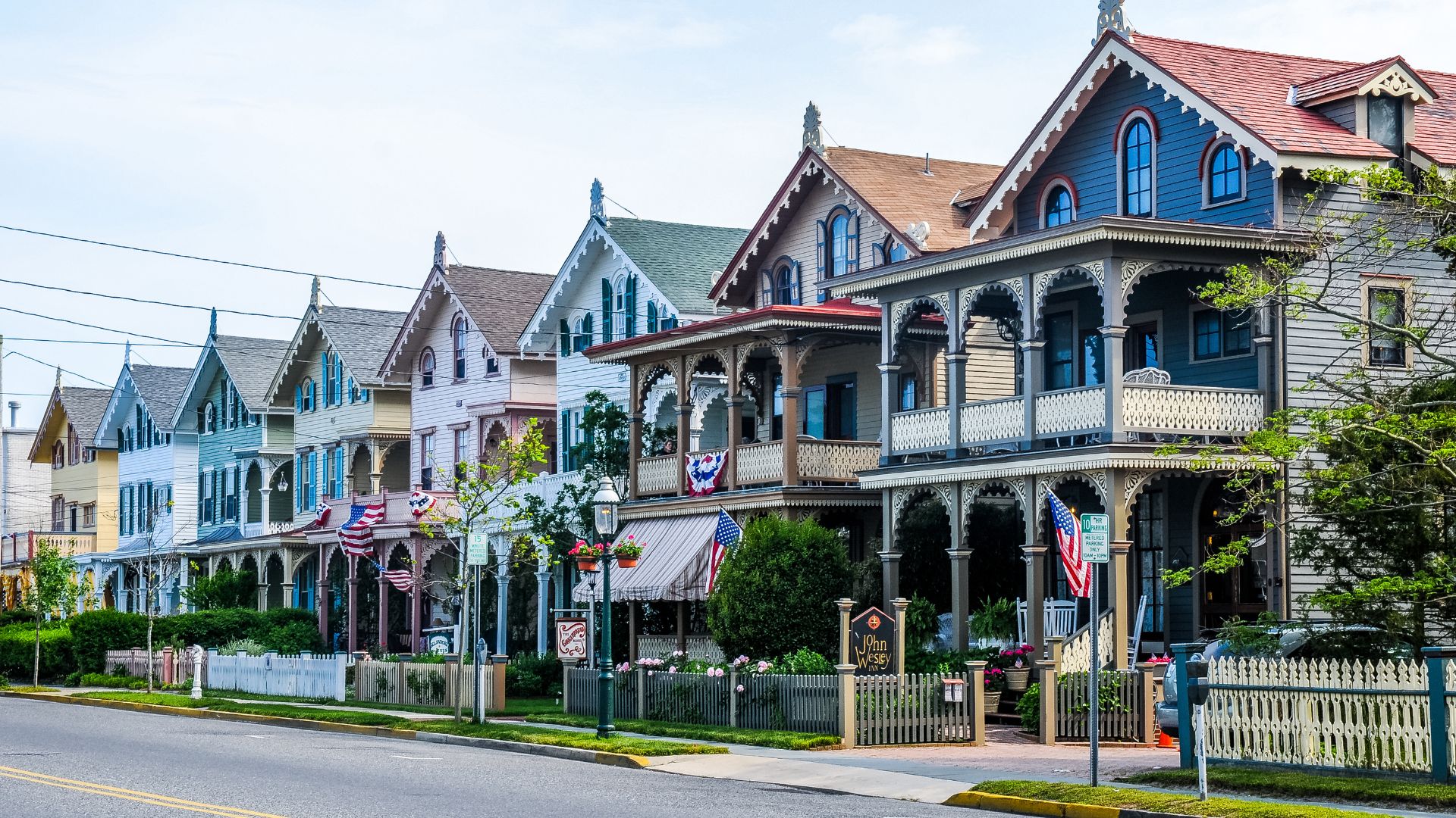 A row of colorful, ornate Victorian houses with intricate details and porches, lining a street under a cloudy sky in Cape May, New Jersey. The houses display various architectural styles and are adorned with flags and fencing.