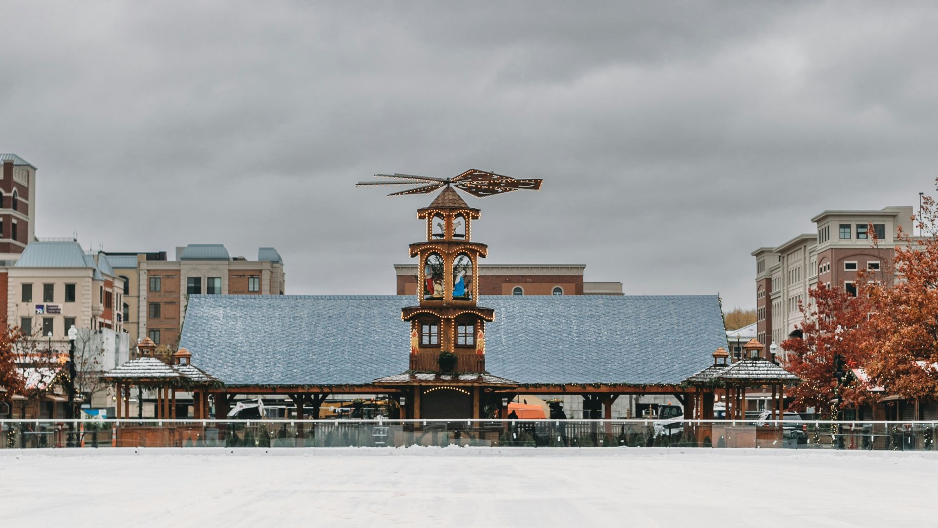 A wide shot of the Carmel Christkindlmarkt in winter, featuring a large, snow-covered ice rink in the foreground and the main market building with a tall, ornate Christmas pyramid on its roof in the background, under a grey, overcast sky. The market building is surrounded by smaller structures, and buildings are visible in the distance.