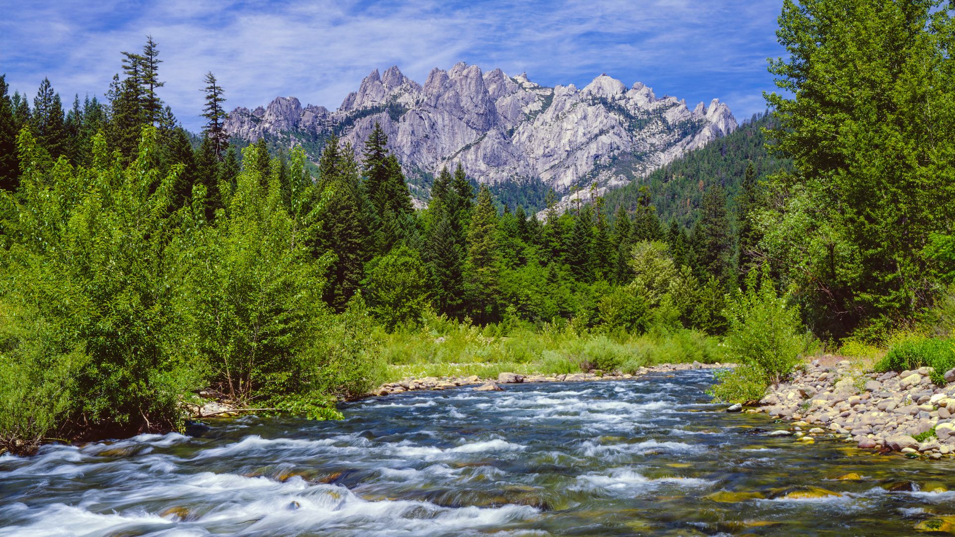 A vibrant image shows a clear river flowing through a lush green forest with towering, rugged granite mountains, known as Castle Crags, in the background under a blue sky.