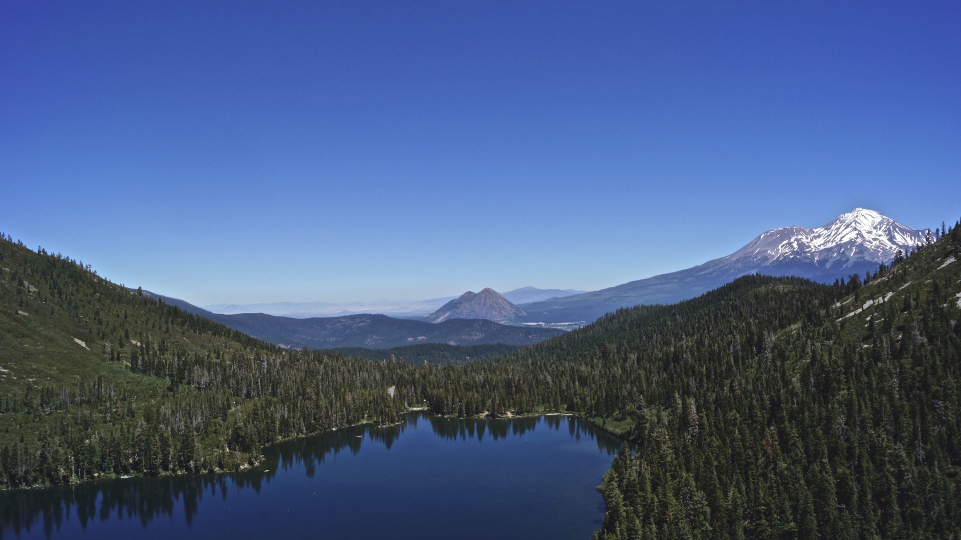 A serene blue mountain lake, Castle Lake, surrounded by dense green pine forests, with the snow-capped peak of Mount Shasta rising majestically in the distance under a clear blue sky.