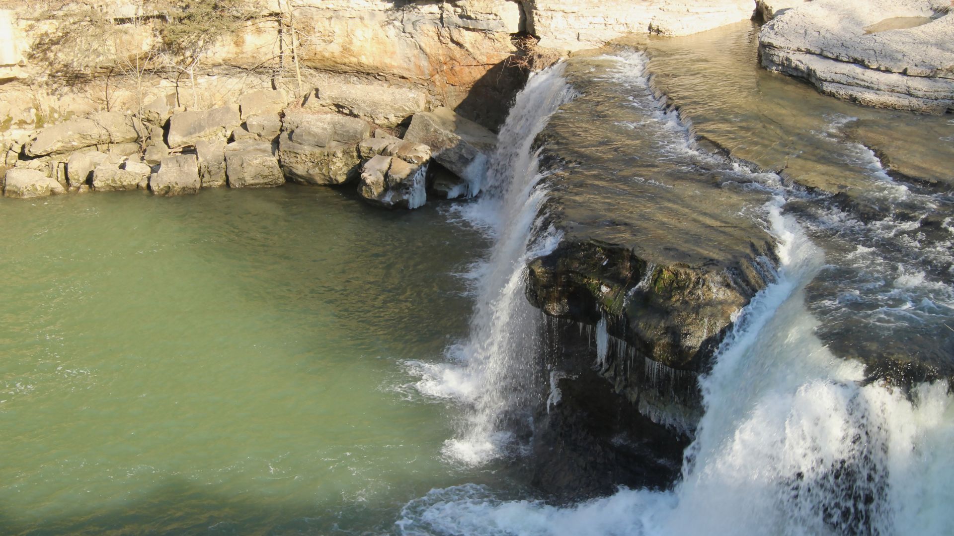 A wide shot of Cataract Falls, showing a multi-tiered waterfall cascading over rocky ledges into a green-tinted pool of water below, with rocky cliffs visible on the left side.