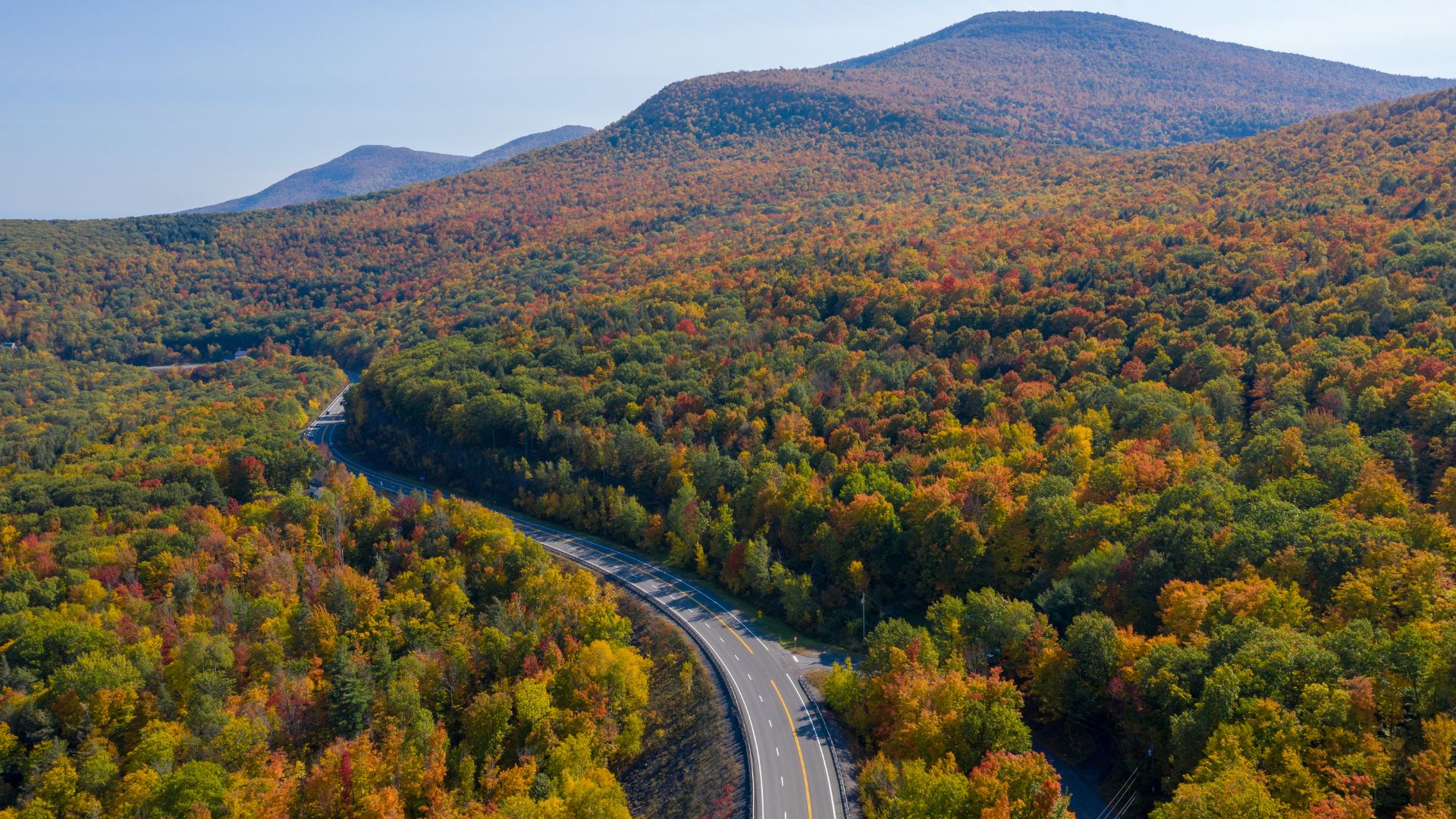 An aerial view shows a winding road cutting through a densely forested mountainous landscape ablaze with vibrant autumn colors of orange, red, and yellow foliage.