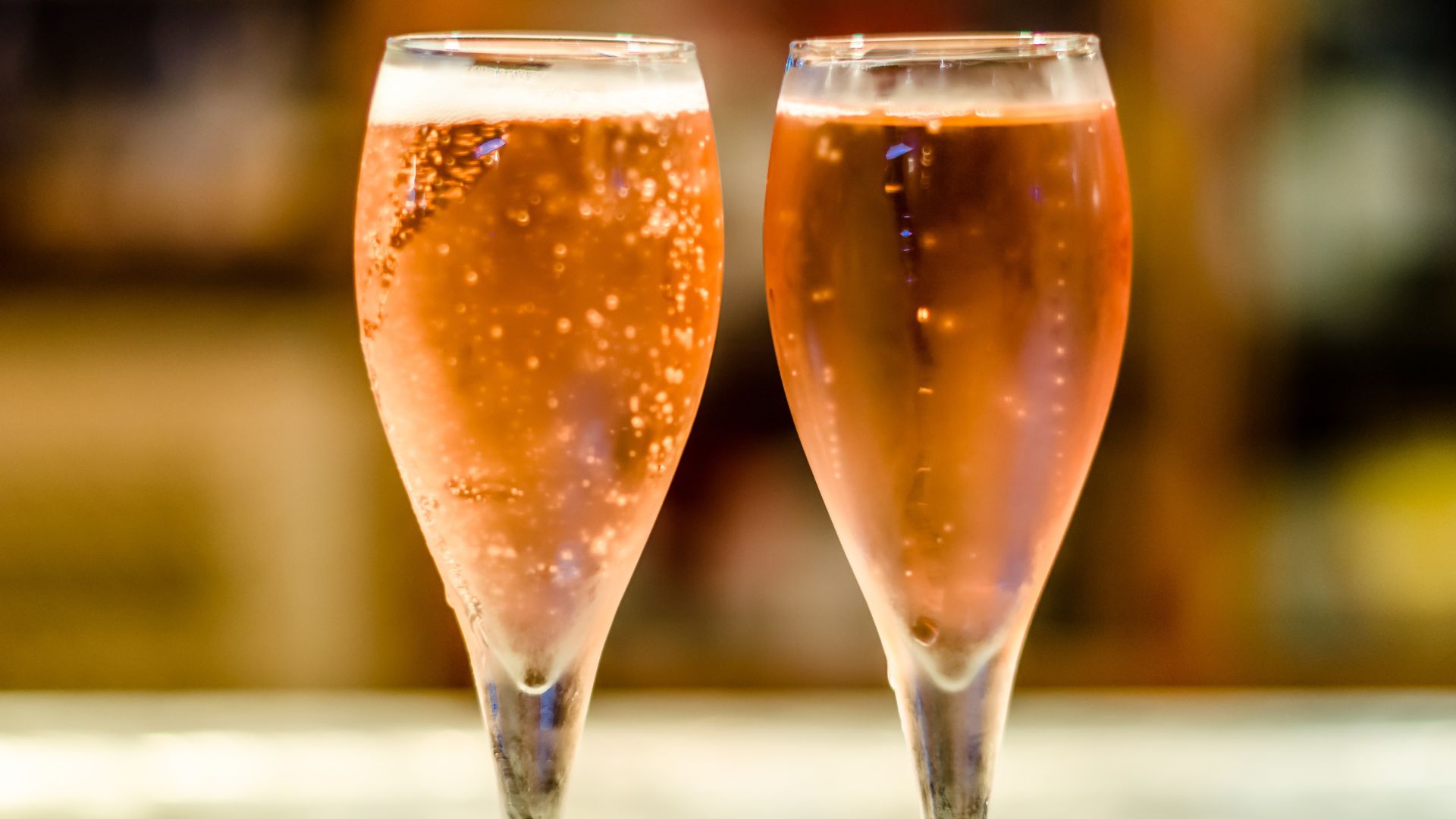 Two flute glasses filled with sparkling rosé-colored wine, showing visible bubbles and foam at the top.