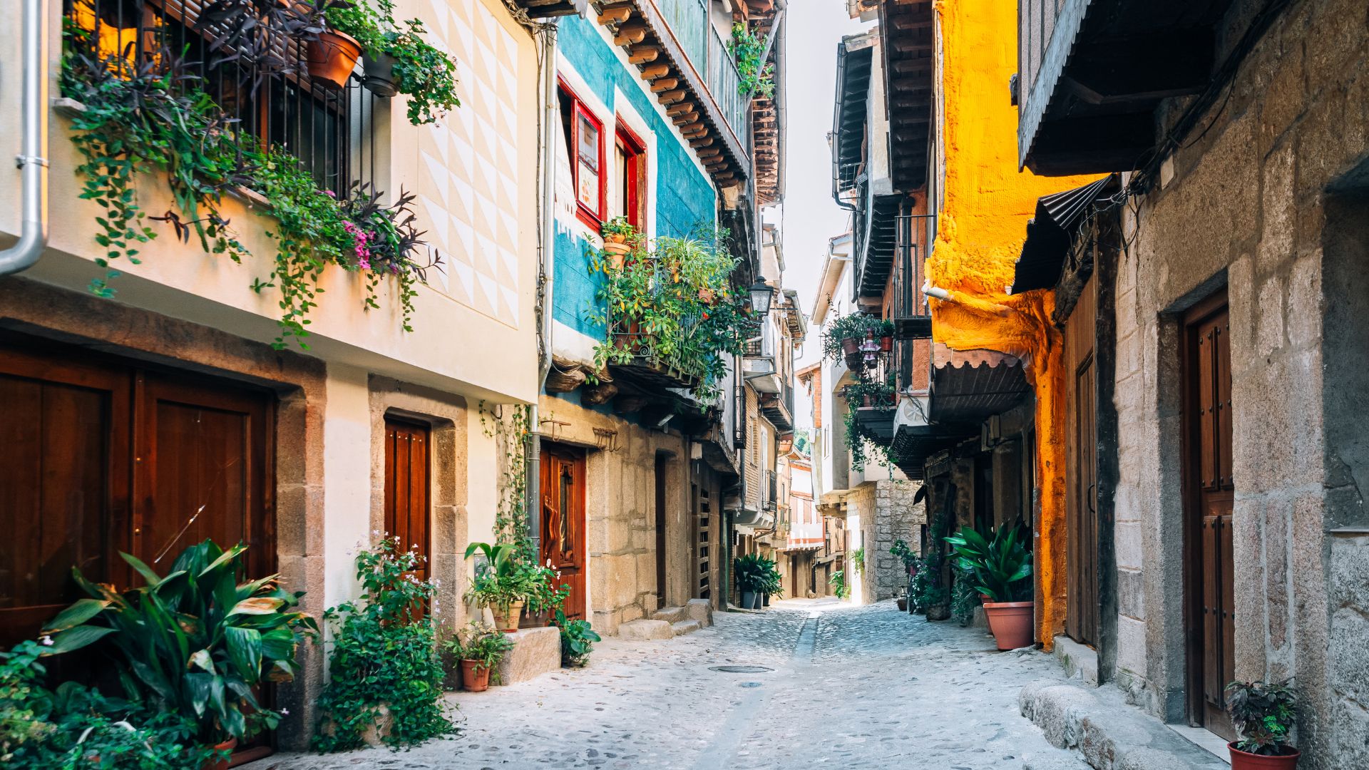 A narrow, cobblestone street winds through a historic Spanish village, flanked by tall buildings with colorful facades, wooden doors, and balconies adorned with lush green plants and vibrant flowers.