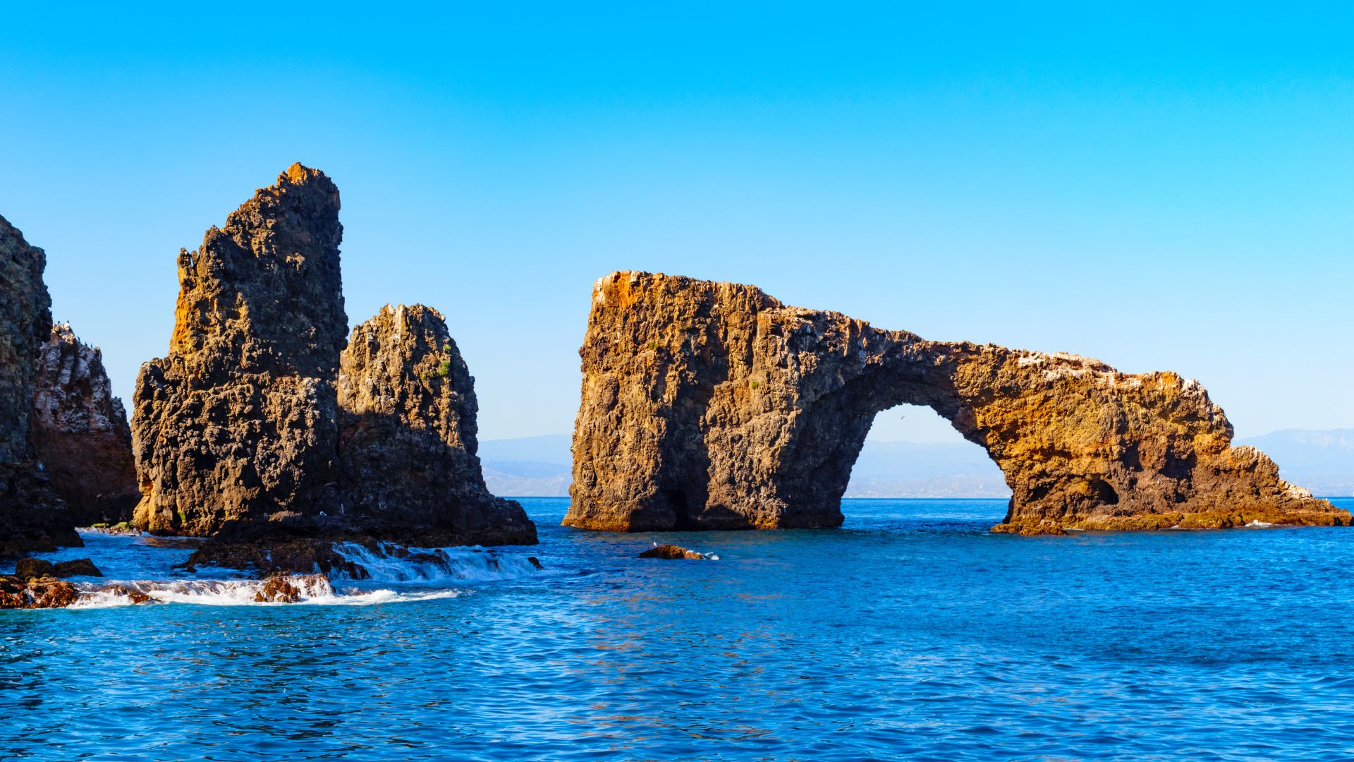 A wide shot of a large, natural rock arch formation, known as Arch Rock, rising from the clear blue ocean under a bright blue sky on Anacapa Island in Channel Islands National Park, California. Smaller rock formations are visible to the left.