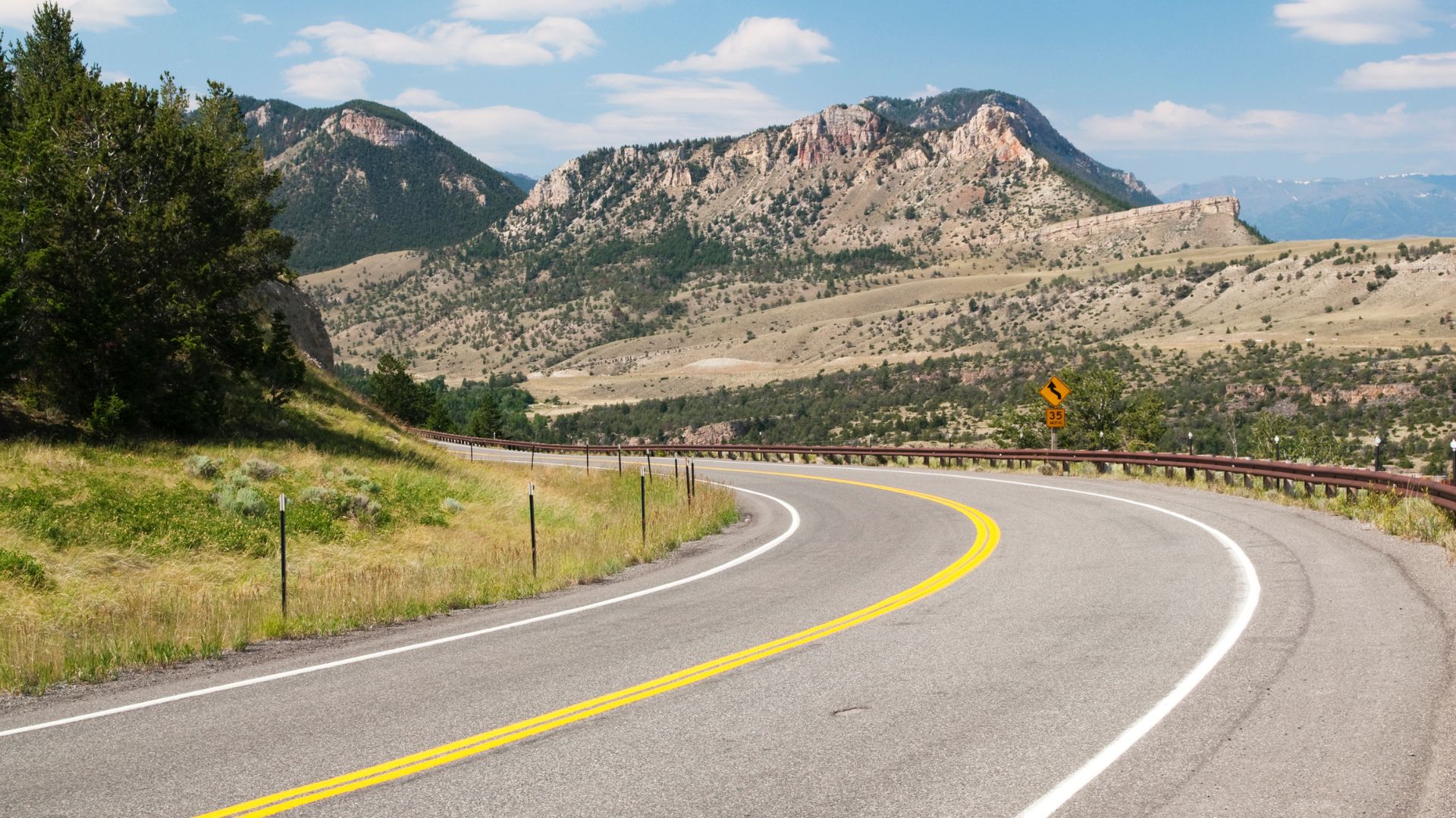 A winding two-lane highway with a double yellow line curves through a mountainous landscape under a partly cloudy sky. The road is flanked by green grass and scattered trees on one side, and a barren, rocky hillside leading to a large mountain range in the distance on the other. A yellow diamond-shaped road sign is visible on the right side of the road.