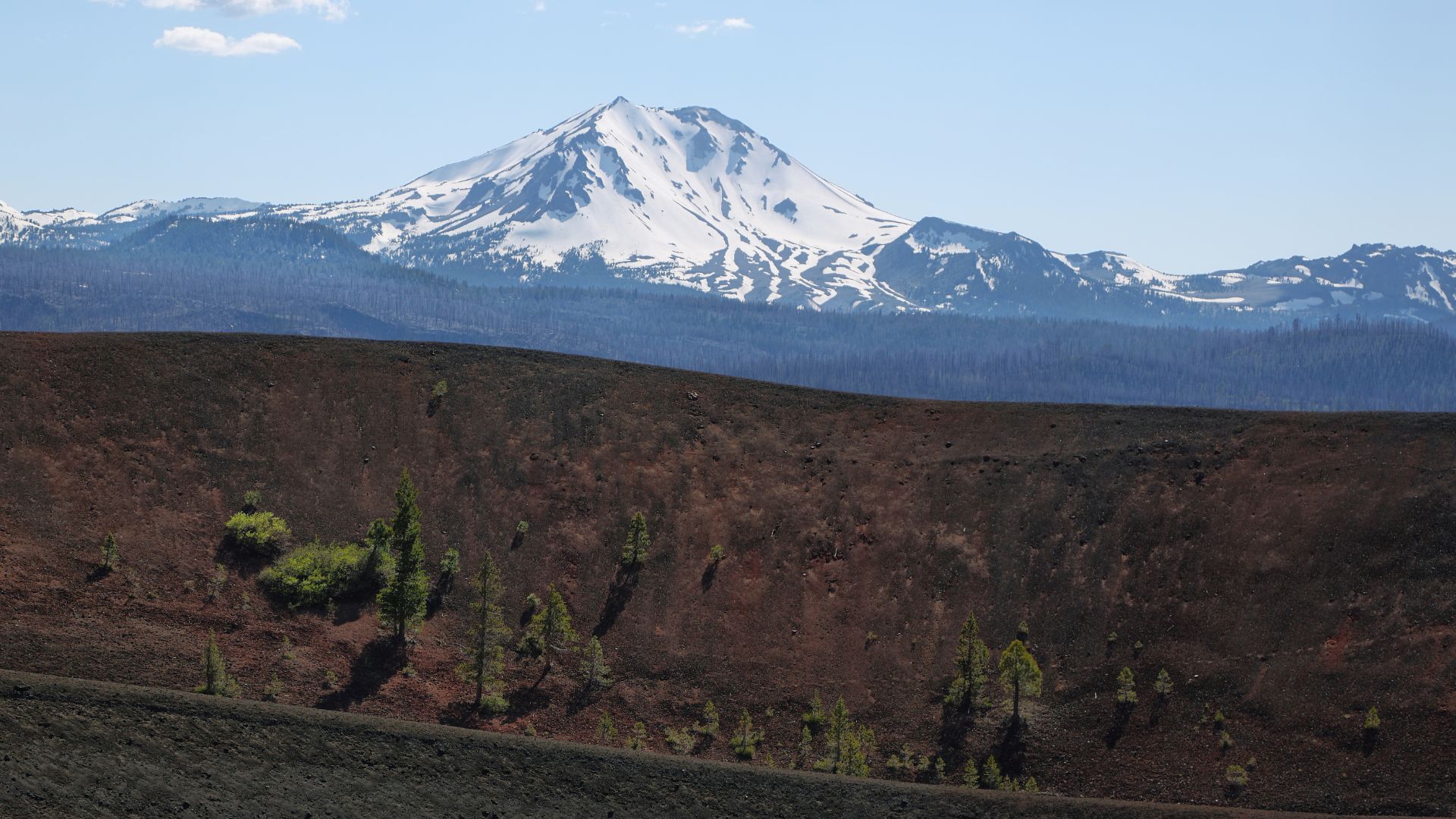 A snow-capped mountain rises in the background behind a dark, barren volcanic landscape with scattered green trees, likely depicting Cinder Cone and its surroundings in Lassen Volcanic National Park, California.