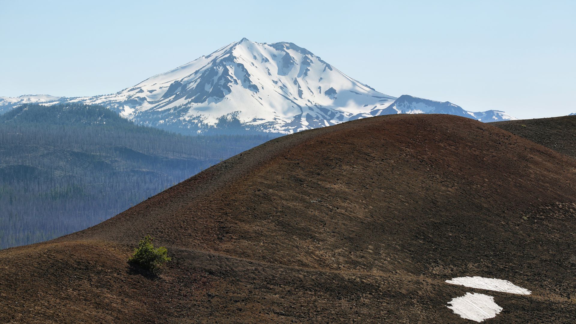 A volcanic landscape featuring a dark, conical cinder cone in the foreground with remnants of snow, and a snow-capped Lassen Peak dominating the background under a clear sky.