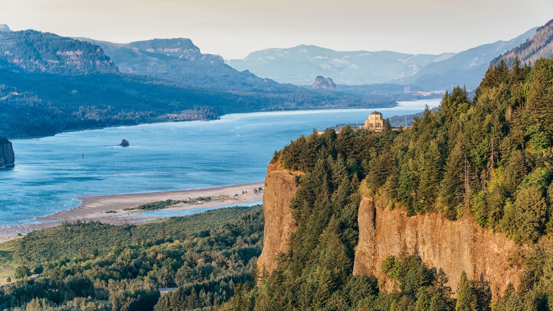 A wide panoramic view of the Columbia River winding through a deep gorge lined with steep, forested cliffs, under a hazy sky, with a historic building (likely Vista House) perched on a prominent cliff edge.