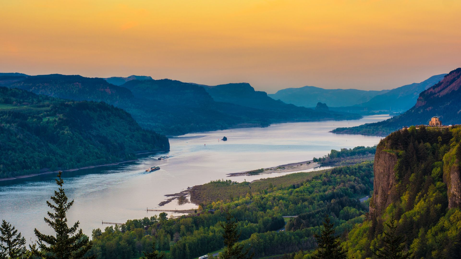 A panoramic aerial view of the Columbia River winding through a forested gorge with mountains in the background under a sunset sky, featuring a prominent cliffside structure on the right.