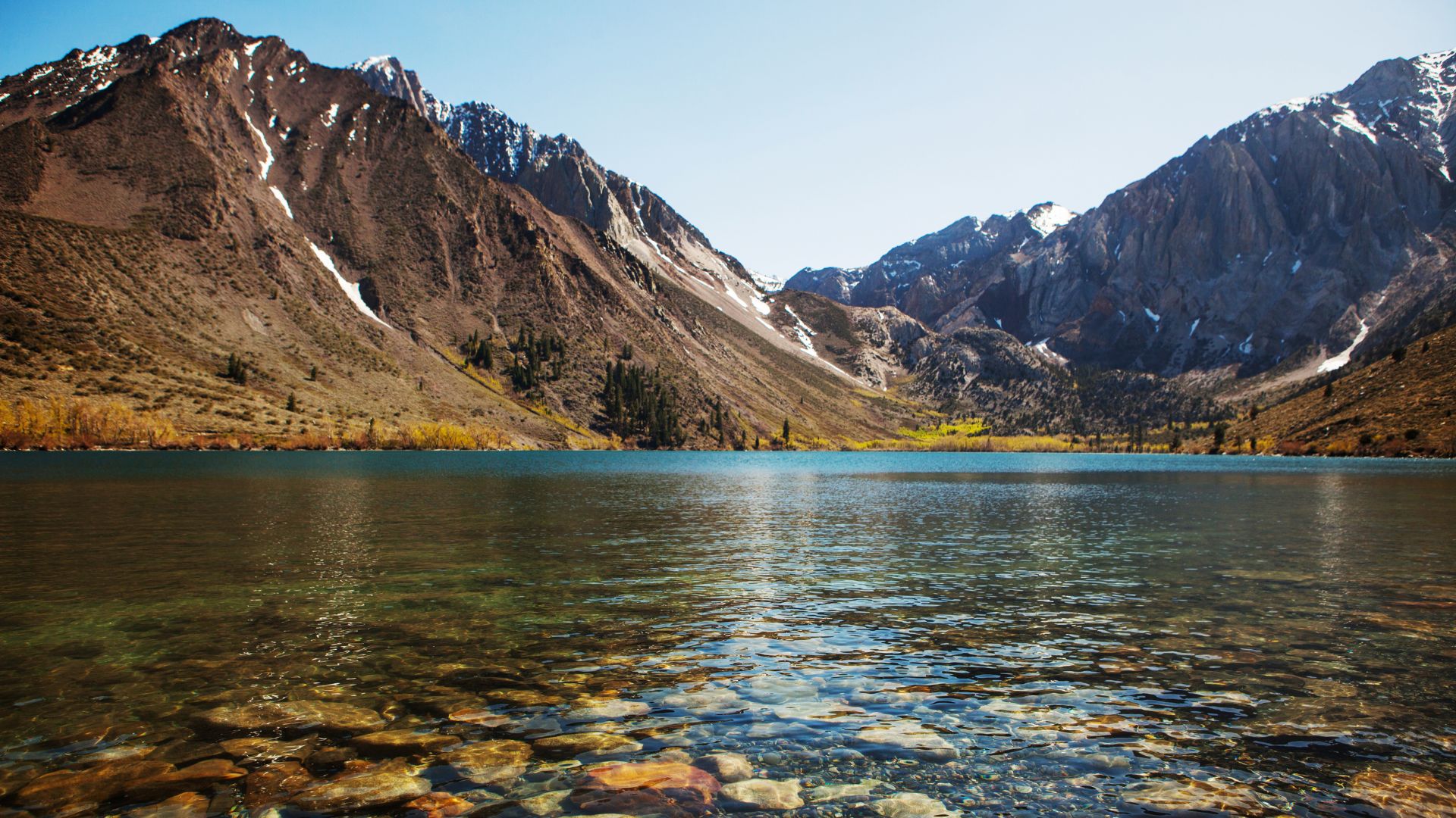 A clear, still lake with visible rocks and pebbles on its shallow bottom, reflecting majestic, rugged mountains under a bright, clear sky. The mountains are largely barren with patches of snow or light vegetation, and a small area of green trees is visible on the far shore.
