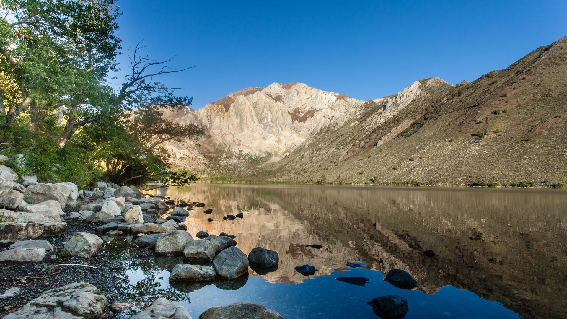 A serene mountain lake, Convict Lake, in California's Eastern Sierra Nevada, with clear blue water reflecting the rugged peaks of the surrounding mountains under a bright blue sky, and a rocky shoreline with scattered vegetation in the foreground.