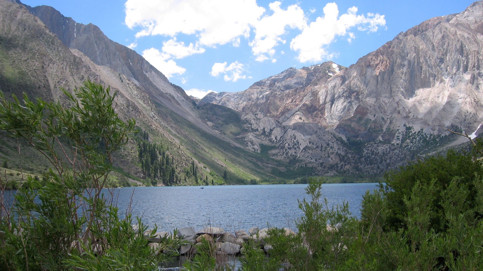 A serene Convict Lake in the Eastern Sierra, California, with towering granite peaks like Laurel Mountain in the background under a blue sky with scattered clouds, framed by green foliage in the foreground.