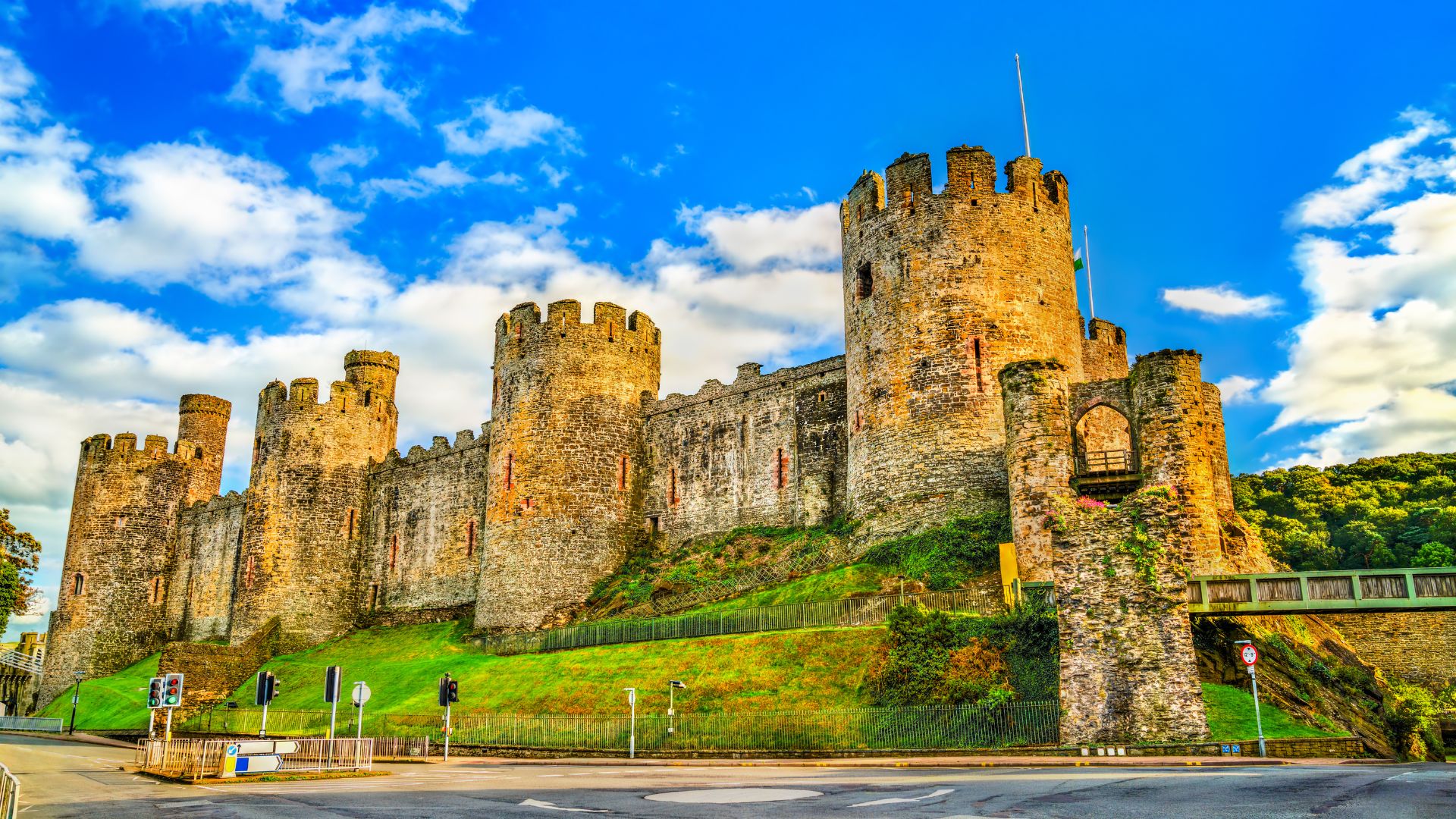 A low-angle view of the impressive, well-preserved Conwy Castle with its high curtain walls and eight lofty towers, set against a bright blue sky with white clouds, overlooking a road and green grassy slopes.