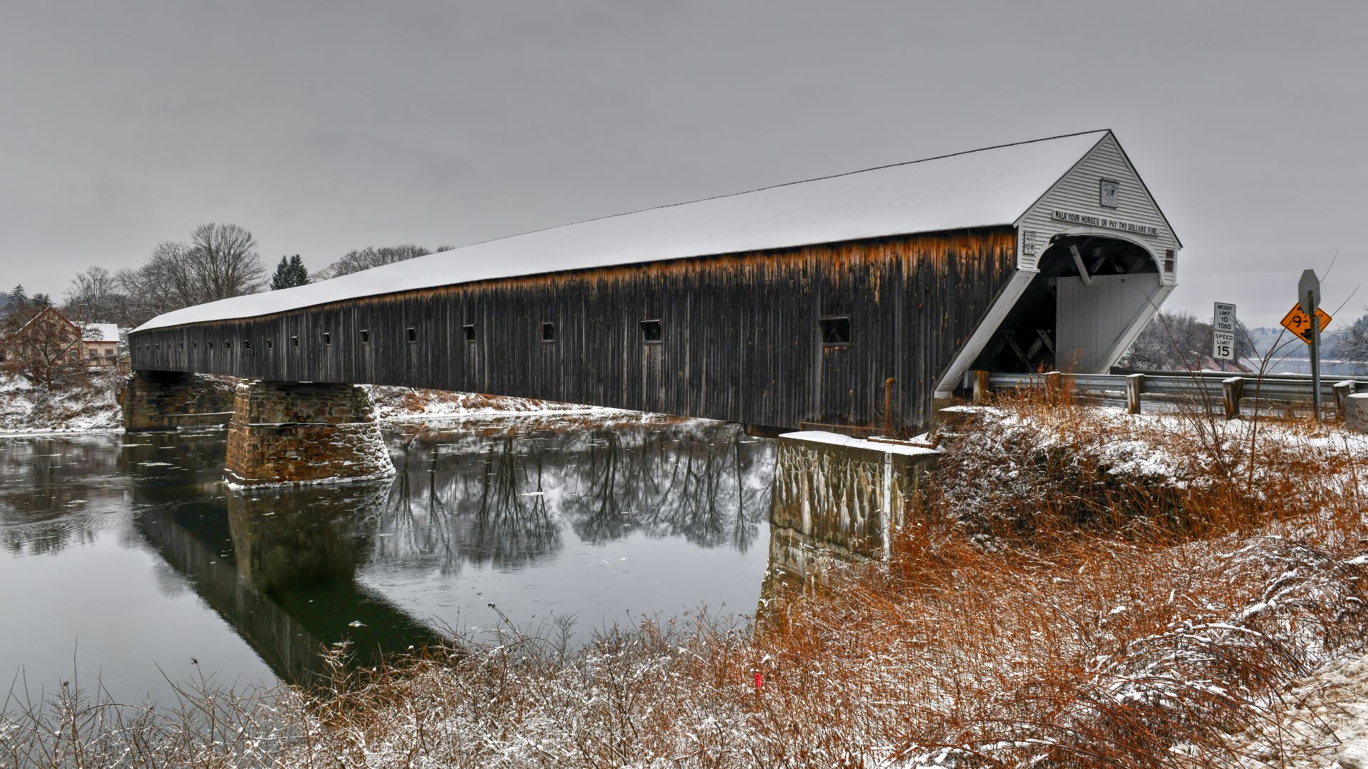 A long, snow-covered wooden covered bridge, the Cornish-Windsor Covered Bridge, extends across a calm river reflecting the winter sky, with snow-dusted banks and bare trees visible in the background.