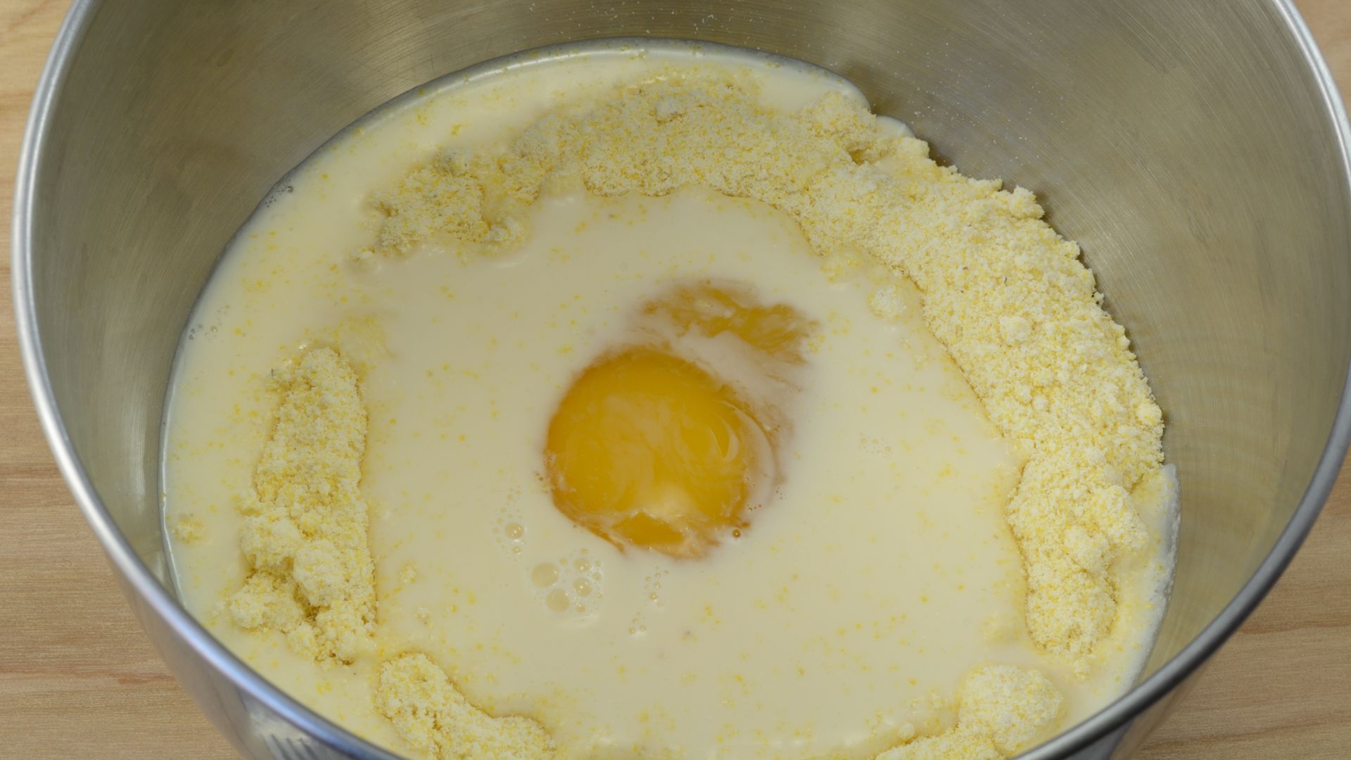 A close-up of a stainless steel mixing bowl containing yellow cornmeal mix, milk, and a raw egg yolk floating in the center.