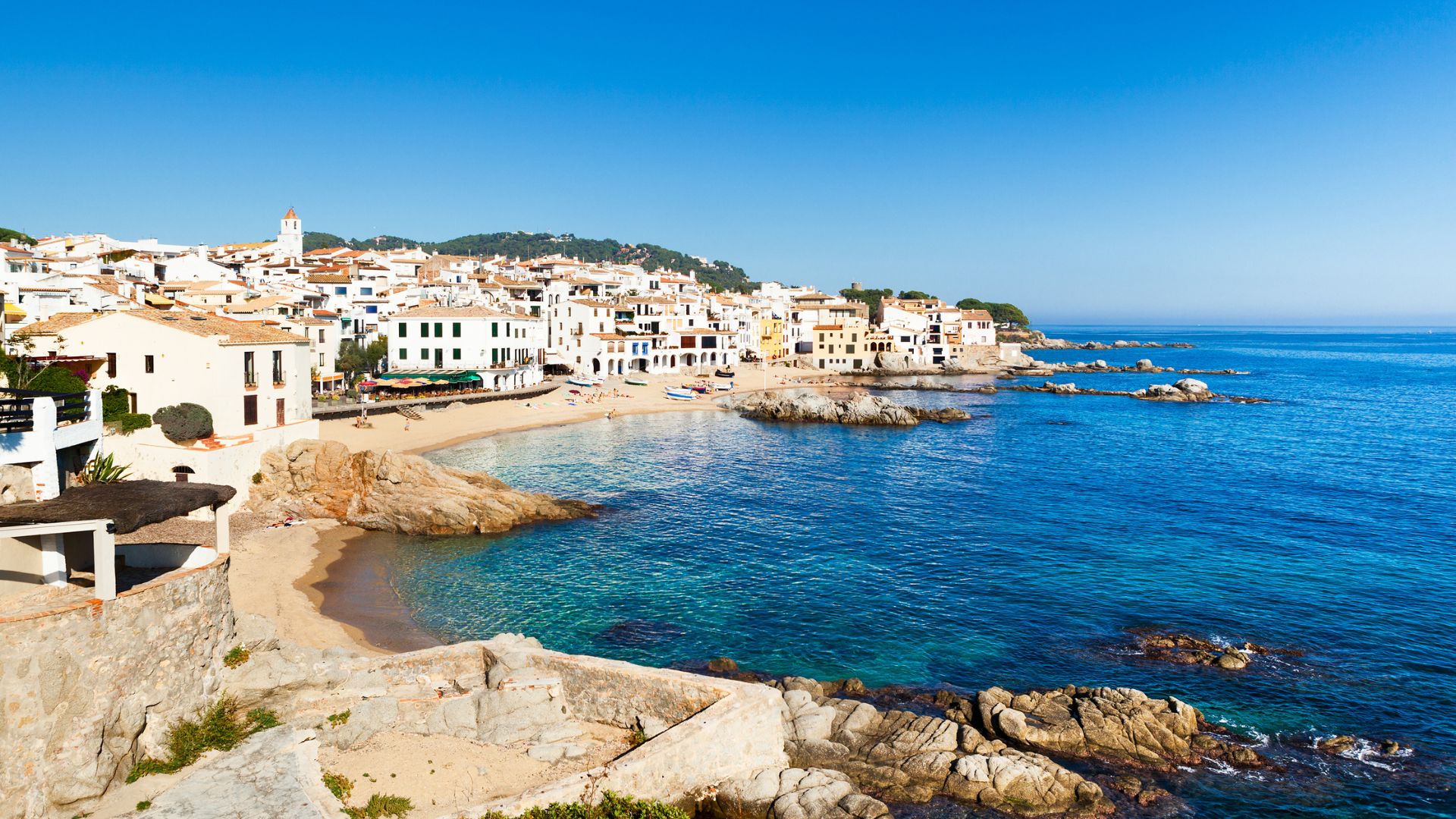 A vibrant, high-angle view of Calella de Palafrugell, a charming Spanish coastal town with whitewashed buildings and red-tiled roofs nestled along a series of small, rocky coves and sandy beaches with clear turquoise waters under a bright blue sky.
