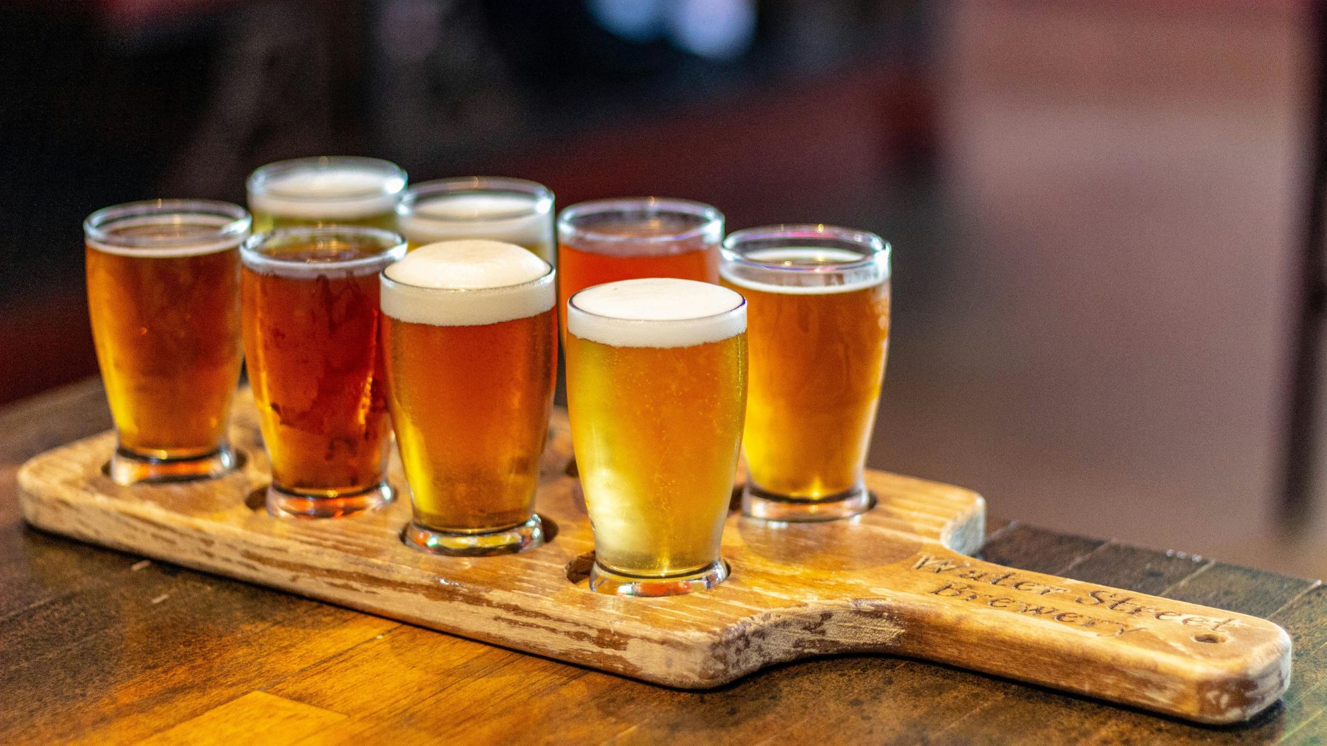 A wooden paddle holds seven small glasses filled with various shades of amber and golden beer, suggesting a craft beer flight for tasting.