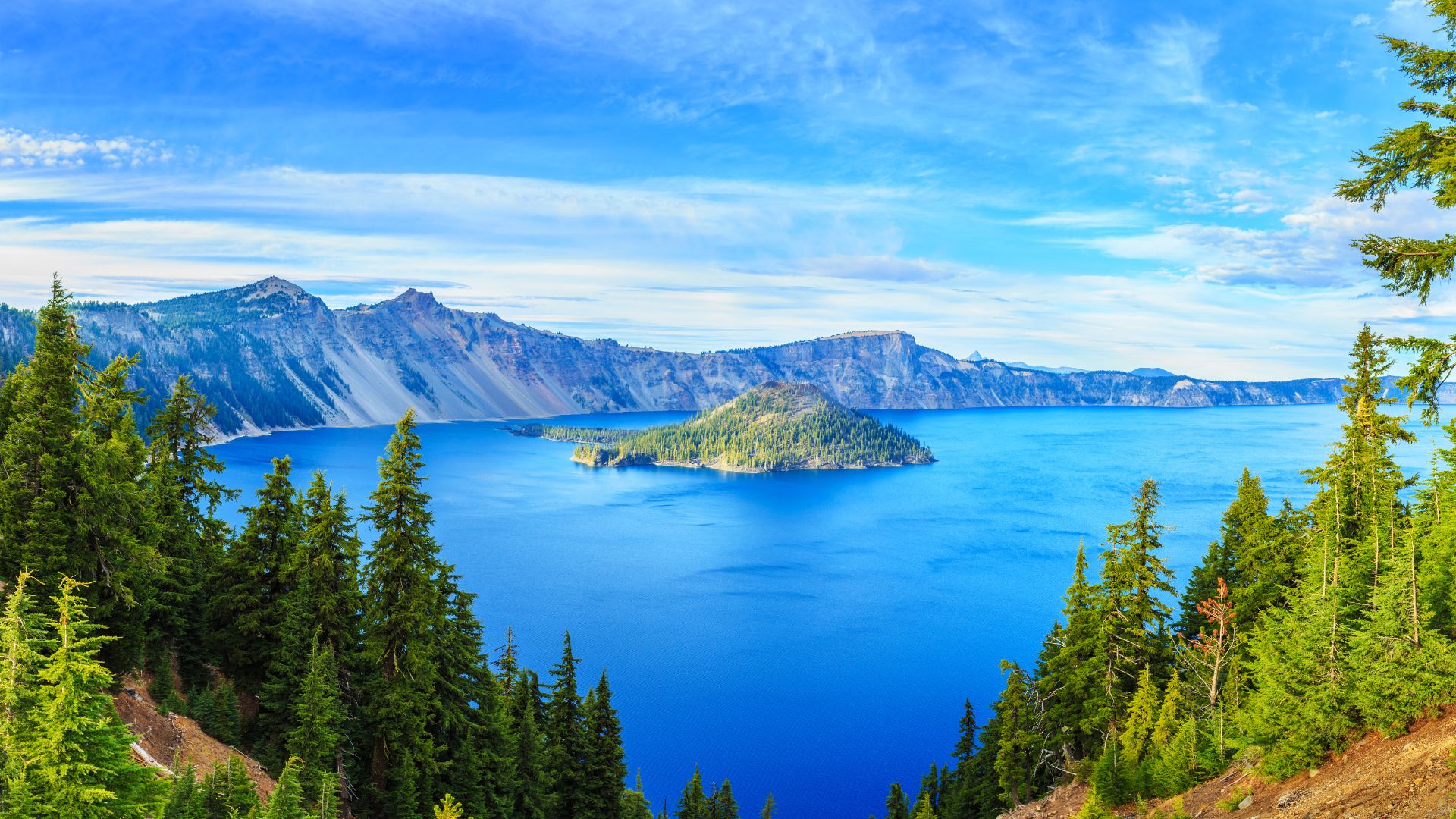 A panoramic view of Crater Lake in Oregon, featuring deep blue water, steep caldera walls, and evergreen trees in the foreground, with Wizard Island visible in the center of the lake.