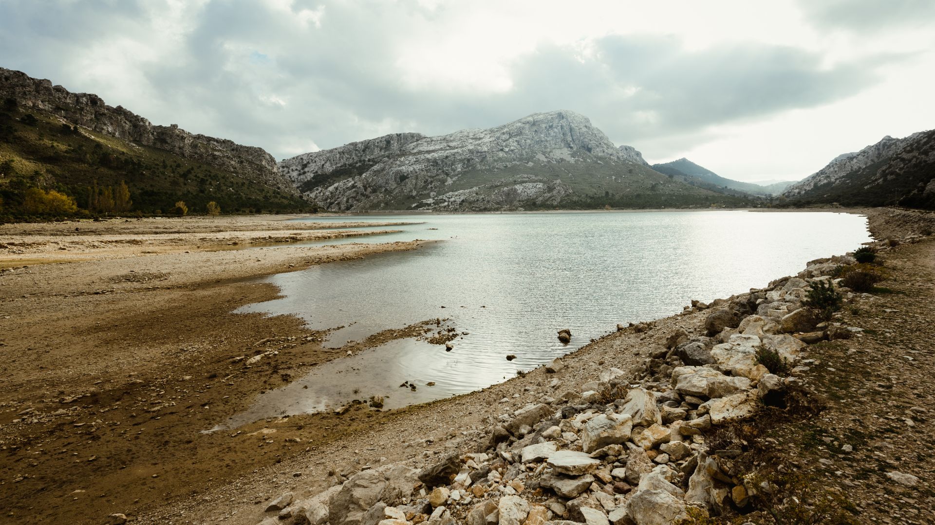 A wide shot of a tranquil lake or reservoir nestled amidst a mountainous landscape under a cloudy sky, with a rocky, arid shore in the foreground.