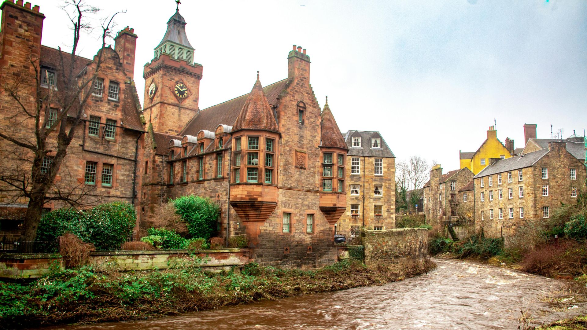A picturesque view of historic stone buildings with red roofs and a prominent clock tower, nestled along the banks of a flowing river in Dean Village, Edinburgh.