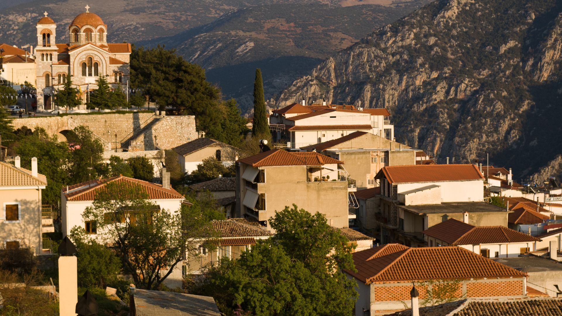 A picturesque view of Delphi, Greece, featuring traditional houses with red-tiled roofs built into a mountainside, leading up to a large church with a domed roof, all set against a backdrop of rugged mountains and a valley below.