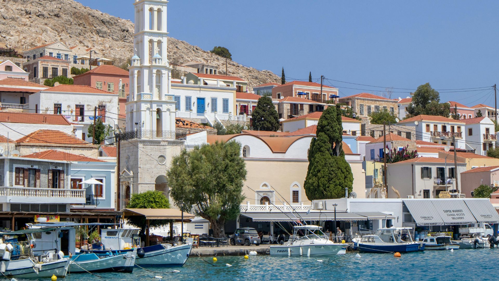 A vibrant image showcasing the harbor of Halki, Greece, with colorful traditional houses cascading down a hillside, a prominent white church tower overlooking the scene, and numerous boats docked in the clear blue waters of the harbor.