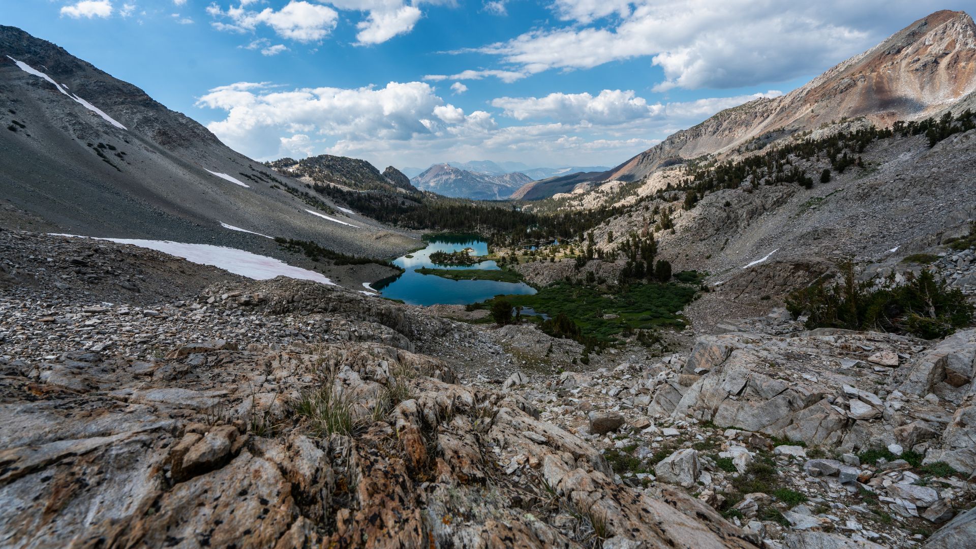 A panoramic view of a mountain pass with snow patches, a series of interconnected blue lakes, and sparse vegetation in a rocky landscape under a partly cloudy sky.