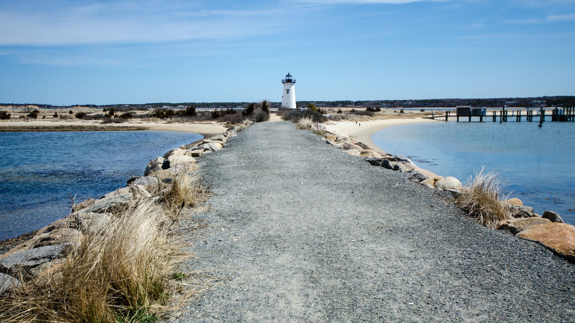 A white lighthouse stands at the end of a stone jetty, extending into a body of water with a sandy beach and distant land visible under a clear sky.