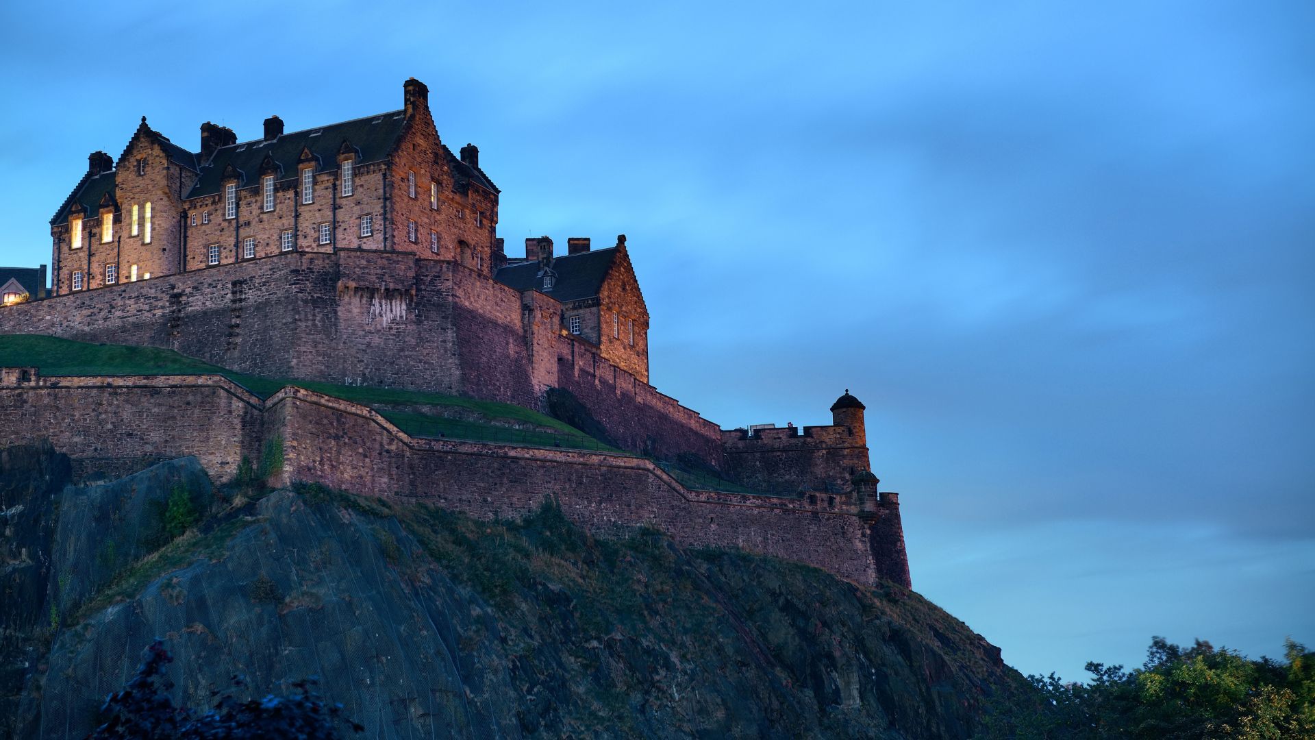 A majestic, illuminated castle sits atop a rocky hill against a twilight blue sky.
