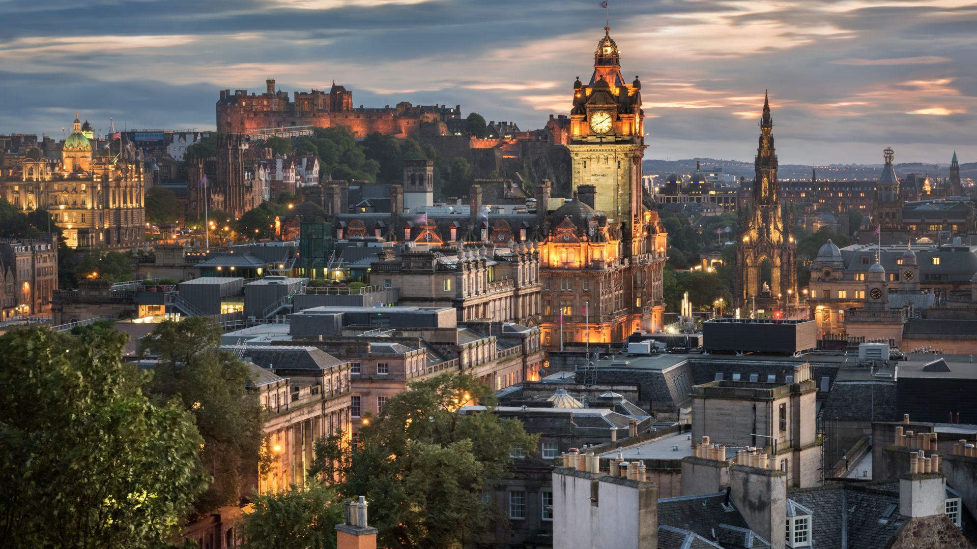 A panoramic view of Edinburgh, Scotland at dusk, showcasing the illuminated Edinburgh Castle perched on Castle Rock, surrounded by historic city buildings and a prominent clock tower.